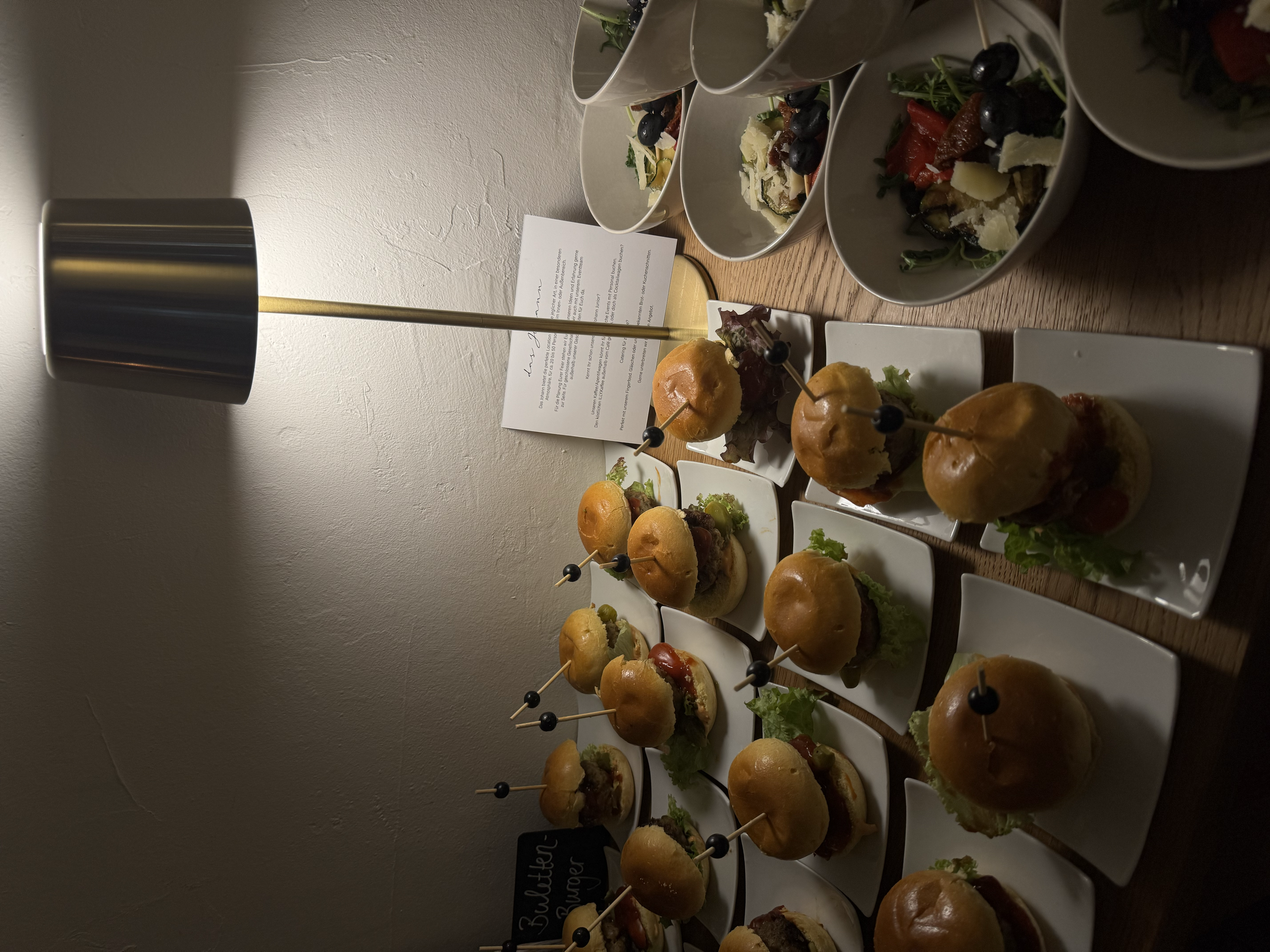 Verschiedene Mini-Burger und Salate auf weißen Tellern, Präsentiert auf einem Holztisch, beleuchtet mit einer modernen Lampe.