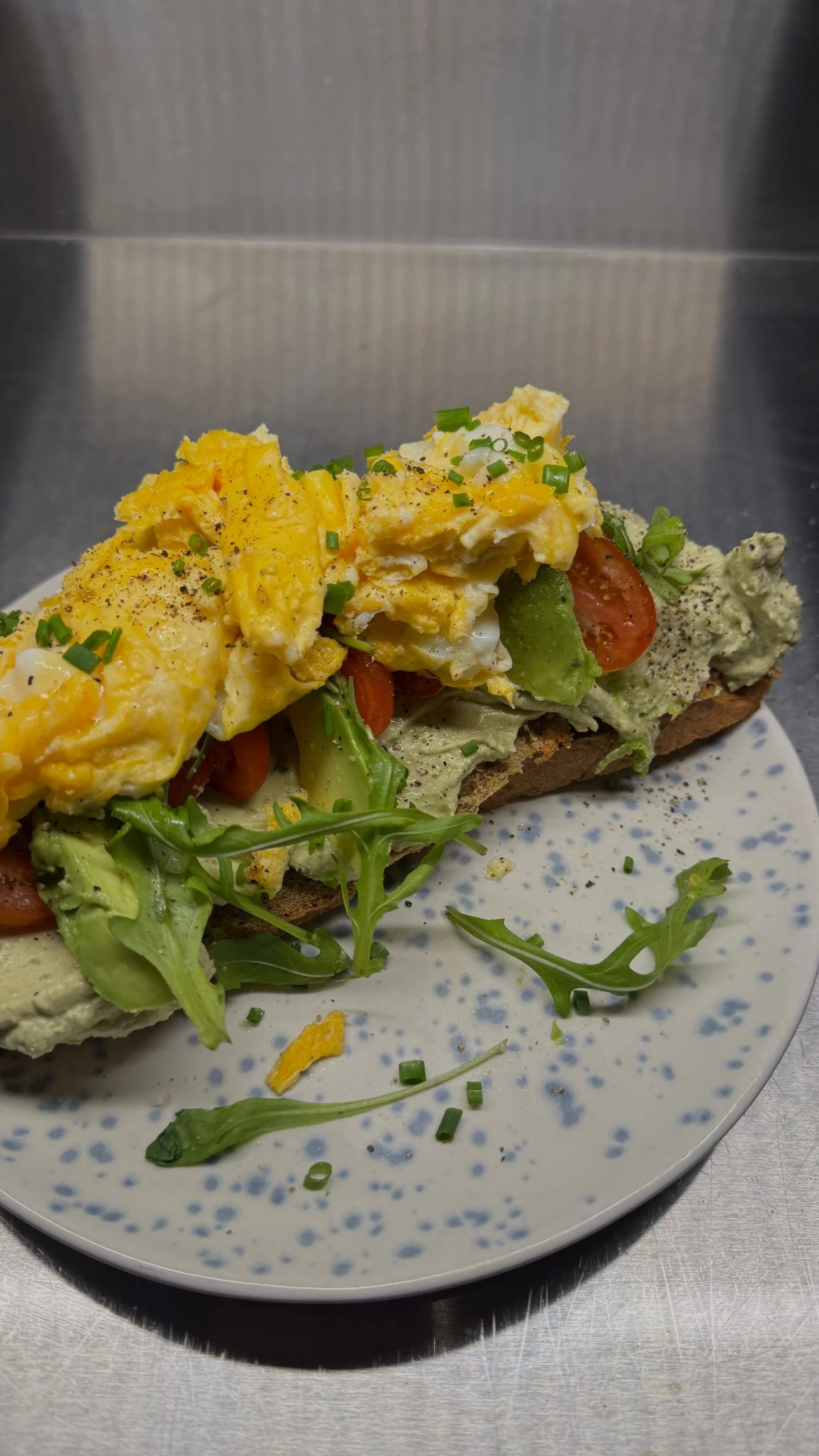 Ein belegtes Brot mit Rührei, Avocado, Tomaten, Salat, und Kräutern auf einem weißen Teller mit blauer Sprenkelung.