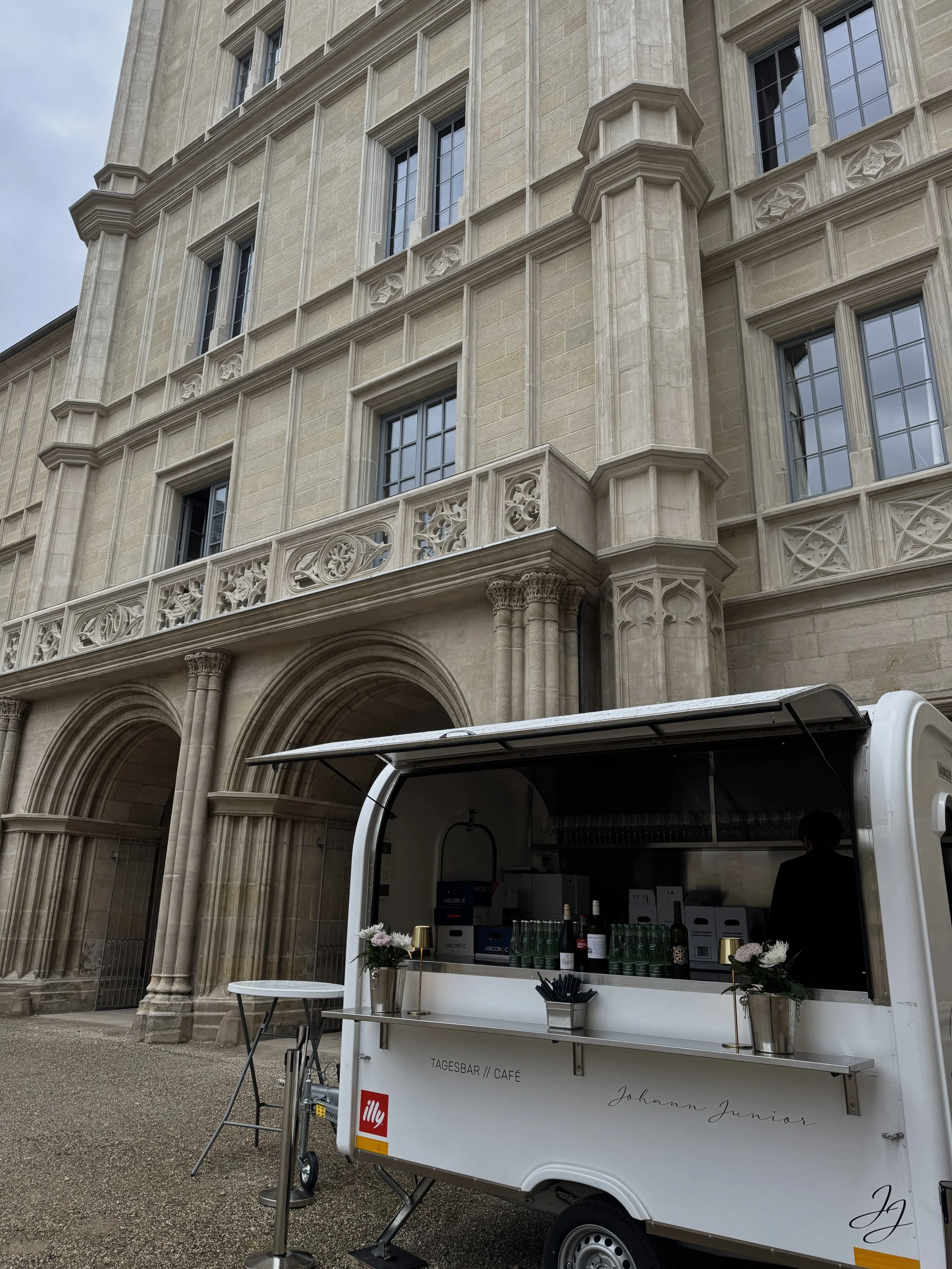 Mobiler Coffee- und Getränkeverkaufsstand im urbanen Außenbereich vor einem historischen Gebäude mit verzierten Fassaden. Der Stand ist weiß mit schwarzen und roten Akzenten und hat Blumen zur Dekoration.