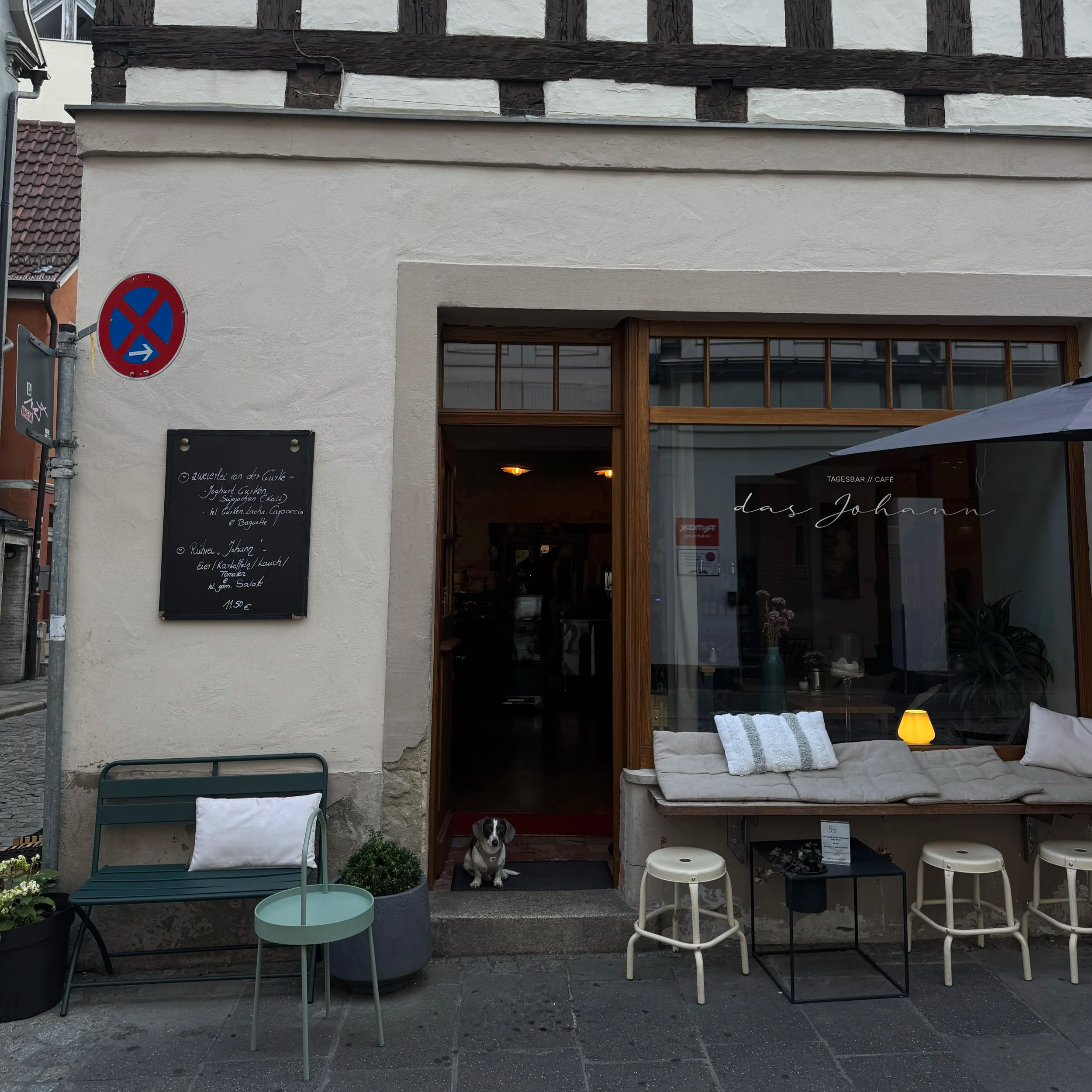 Außenansicht eines Cafés mit Sitzbereich, einem schwarzen Hund in der Türöffnung, mehreren Stühlen, Tischen, Pflanzen und Dekorationen, darunter Laternen und Stoffkissen.