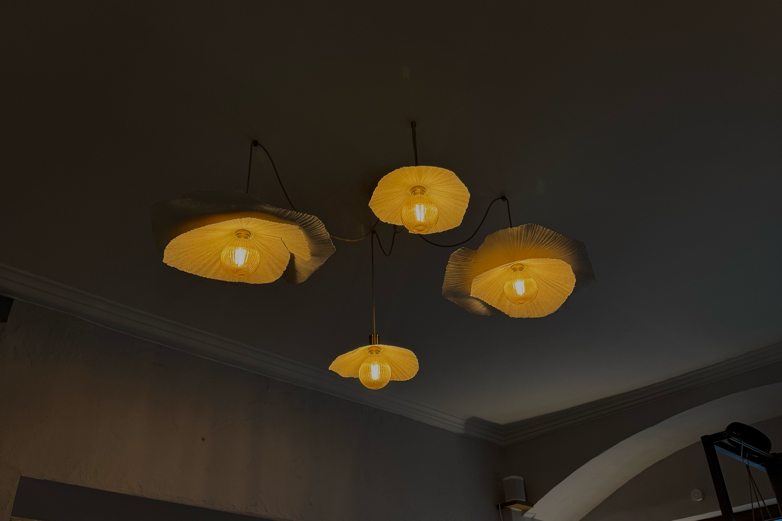 Hängende Lampe mit vier Lampen, die mit gelben, papierartigen Lampenschirmen bedeckt sind, an einem dunklen Deckenhimmel.