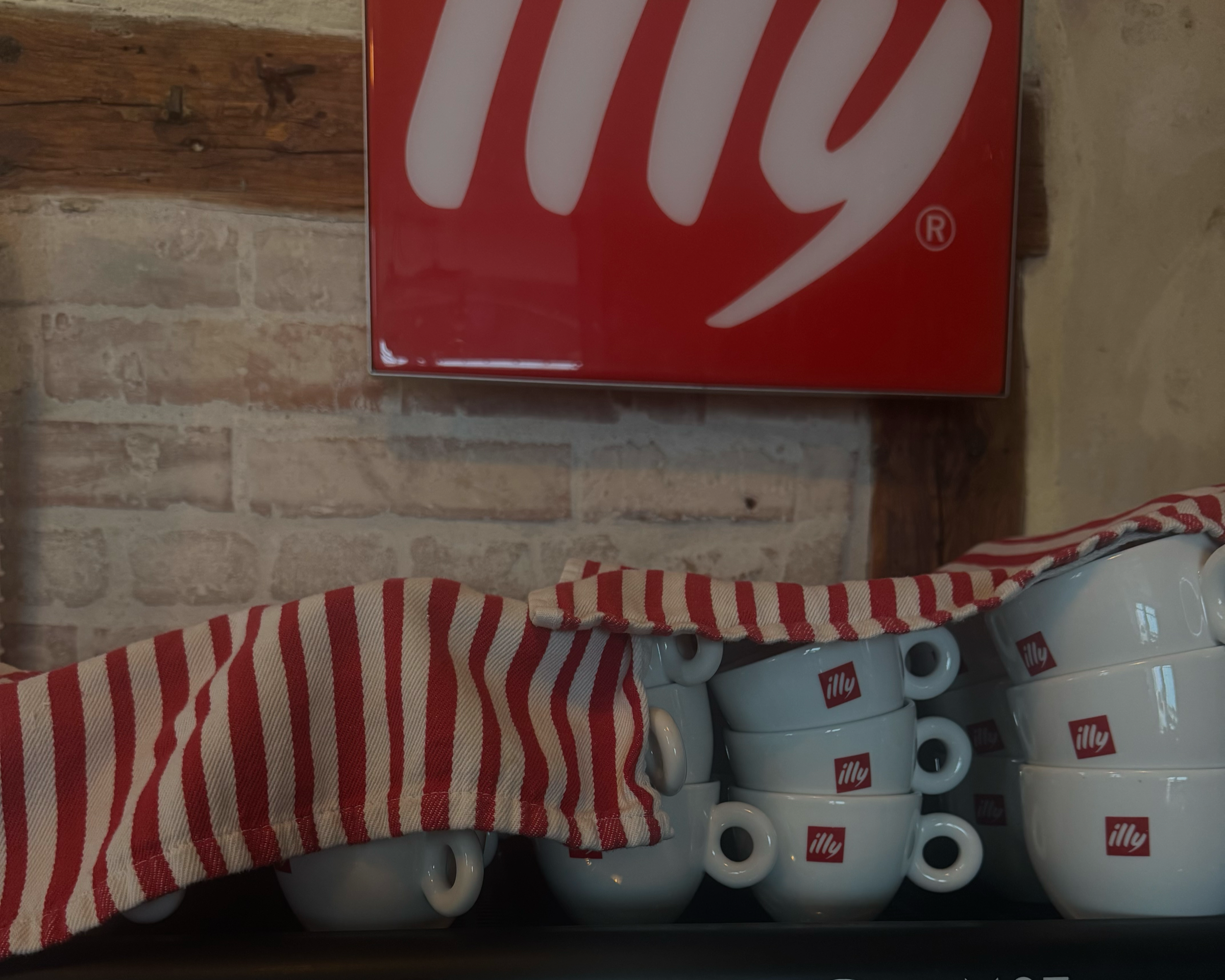 Mehrere weiße Kaffeetassen mit rotem Illy-Logo, übereinander gestapelt, und ein rotes und weiß gestreiftes Küchentuch, auf einem Regal gegen eine Backsteinwand.
