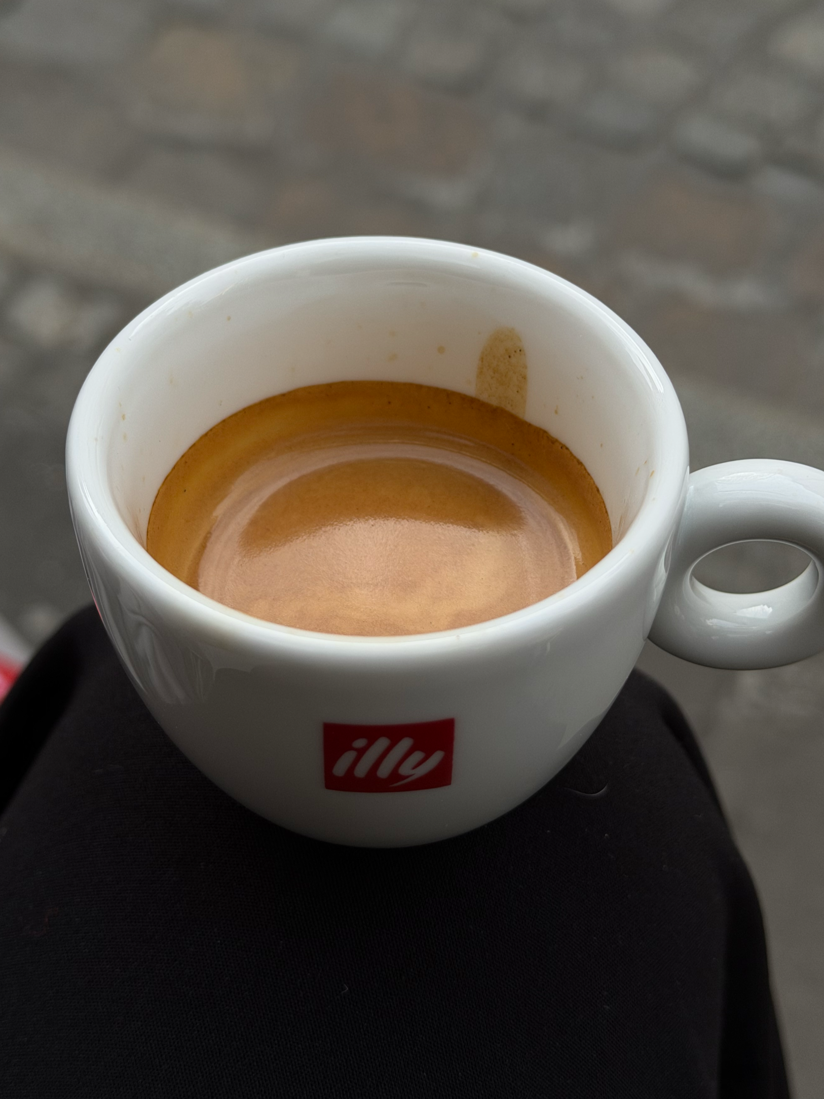 Eine weiße Kaffeetasse mit dem Illy-Logo, gefüllt mit Espresso, auf einem schwarzen Untersetzer, im Hintergrund ein grauer, gepflasterter Boden.