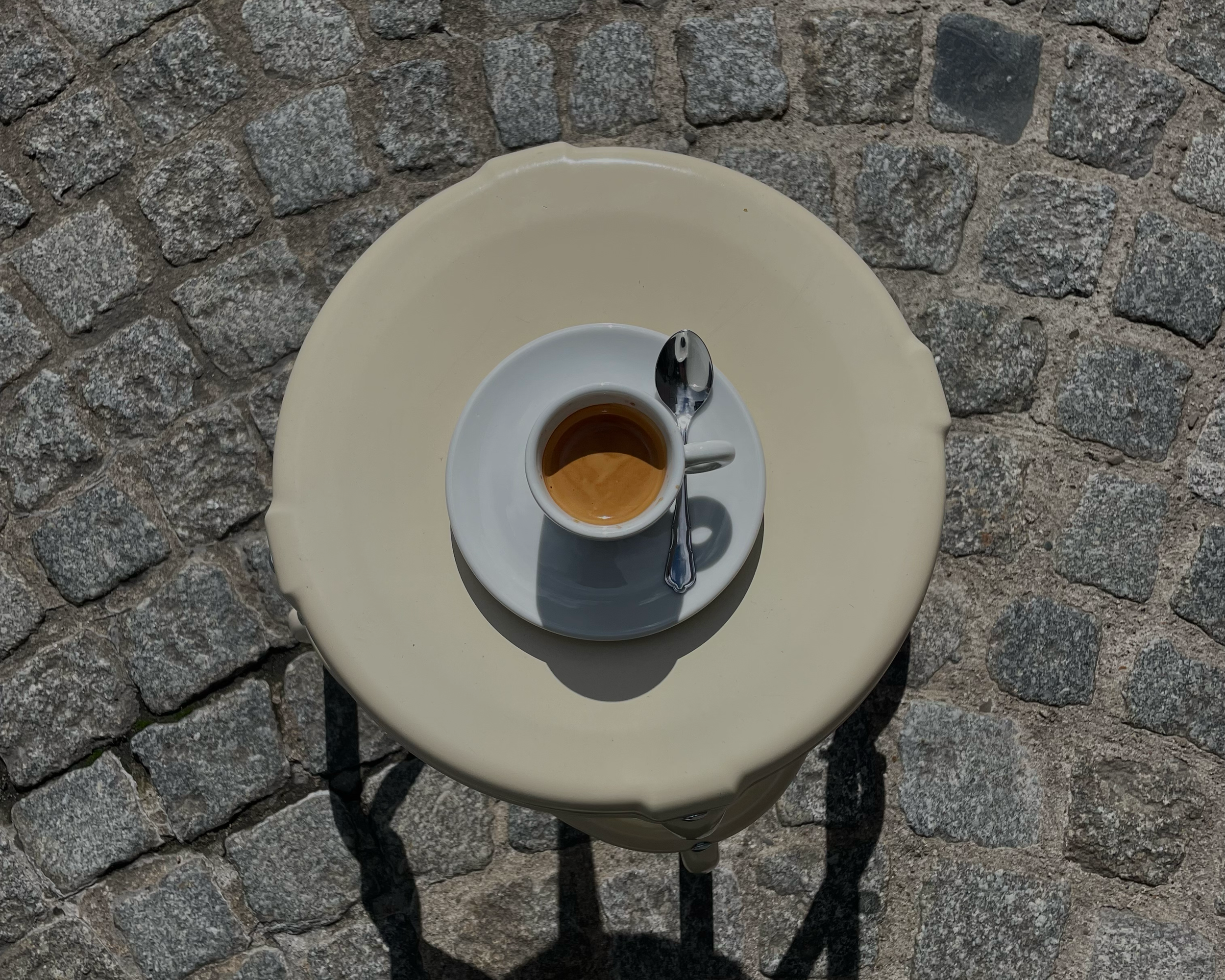 Eine weiße Tischplatte mit einer weißen Kaffeetasse und einem Silberlöffel, im Hintergrund ein Kopfsteinpflasterboden.