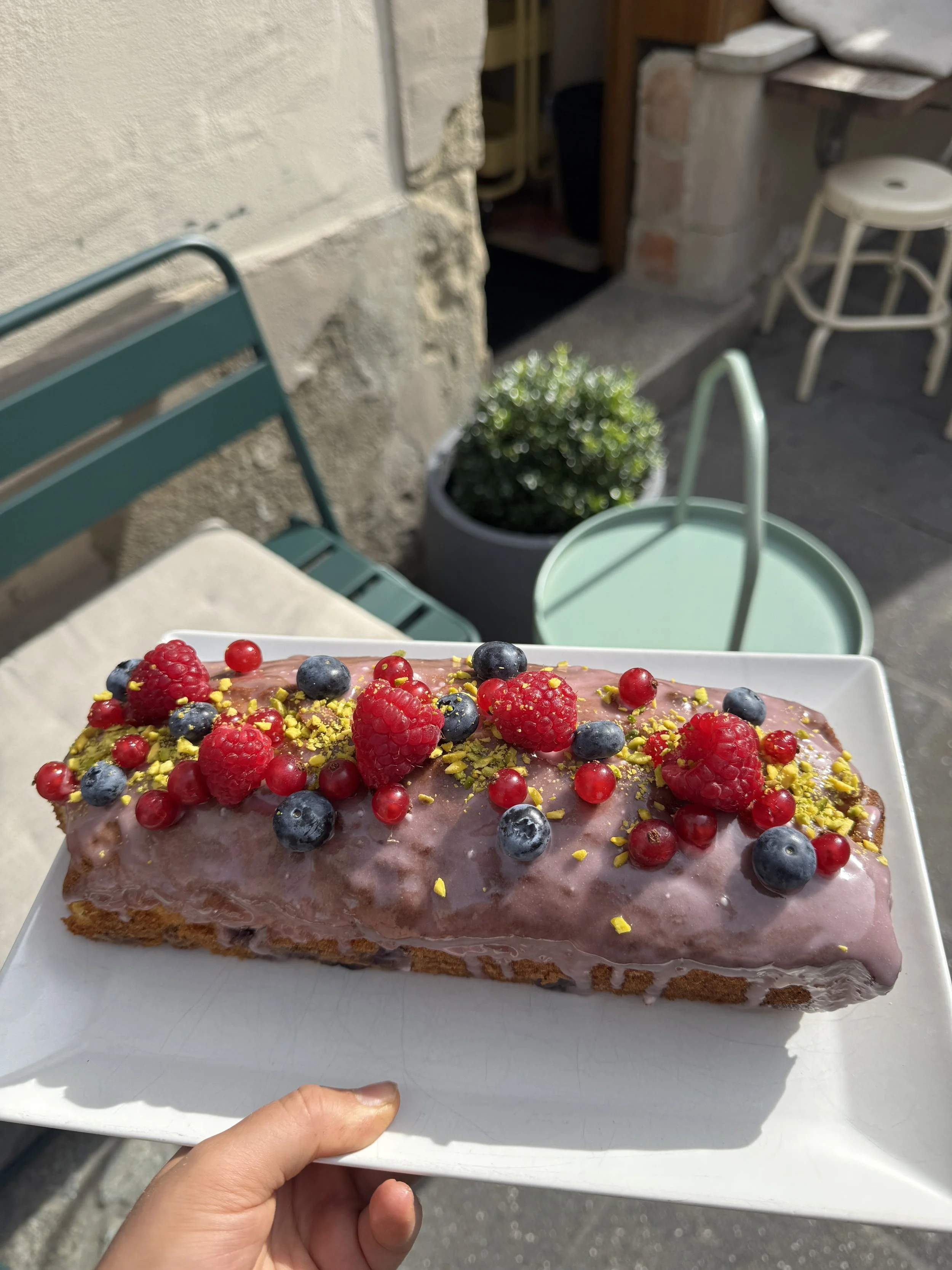Ein Stück Kuchen mit rosa Glasur, verziert mit roten und blauen Beeren sowie grünen Pistazien. Es wird auf einem weißen Teller gehalten.