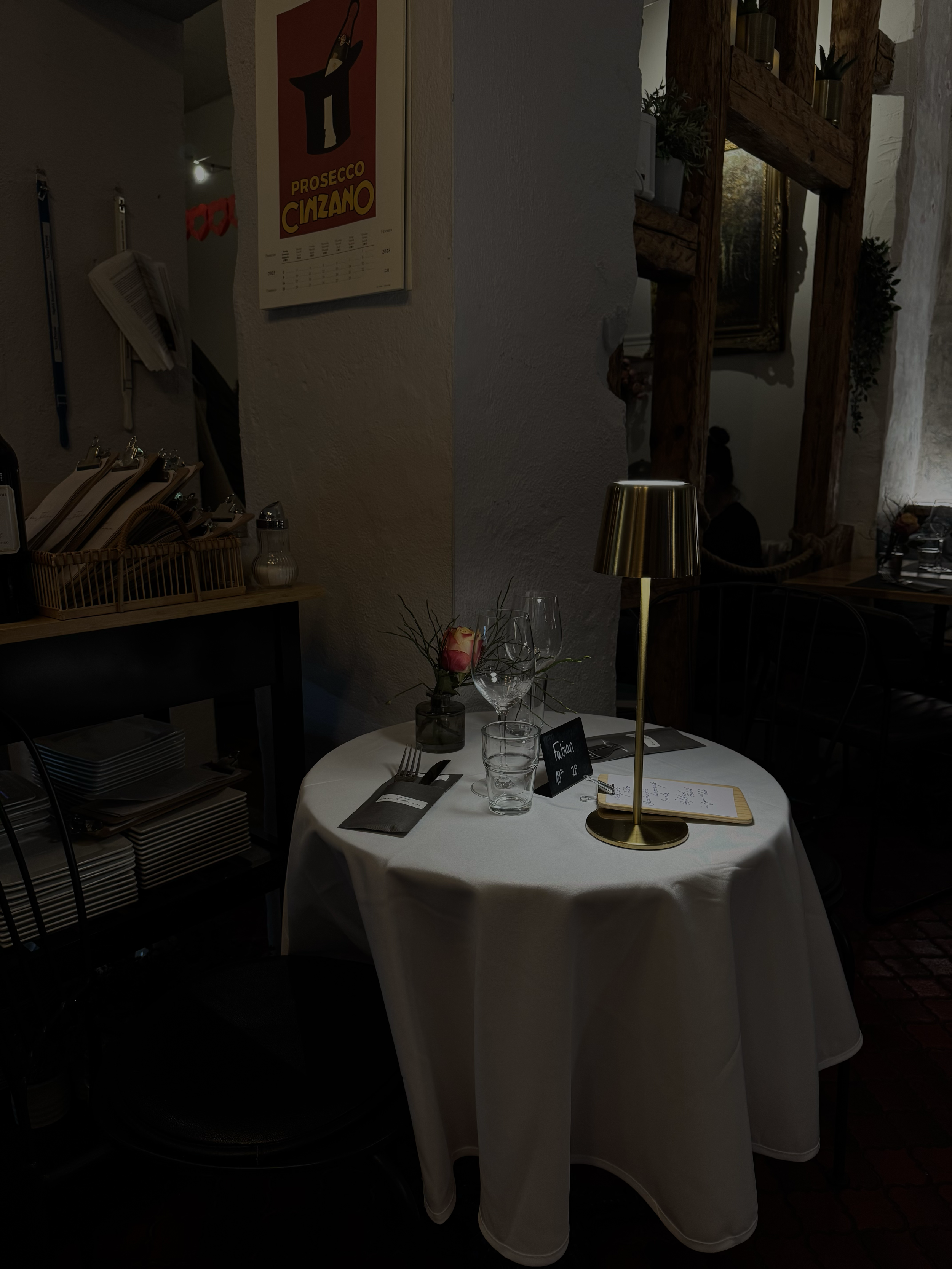 Ein gedeckter Esstisch mit weißen Tischtuch, einem Kerzenglas, einer kleinen Vase mit Rose, einem Notizbuch, Menükarte, Wasser- und Weingläser, in einem rustikalen Restaurant mit dunklen Holzmöbeln und Dekorationen.