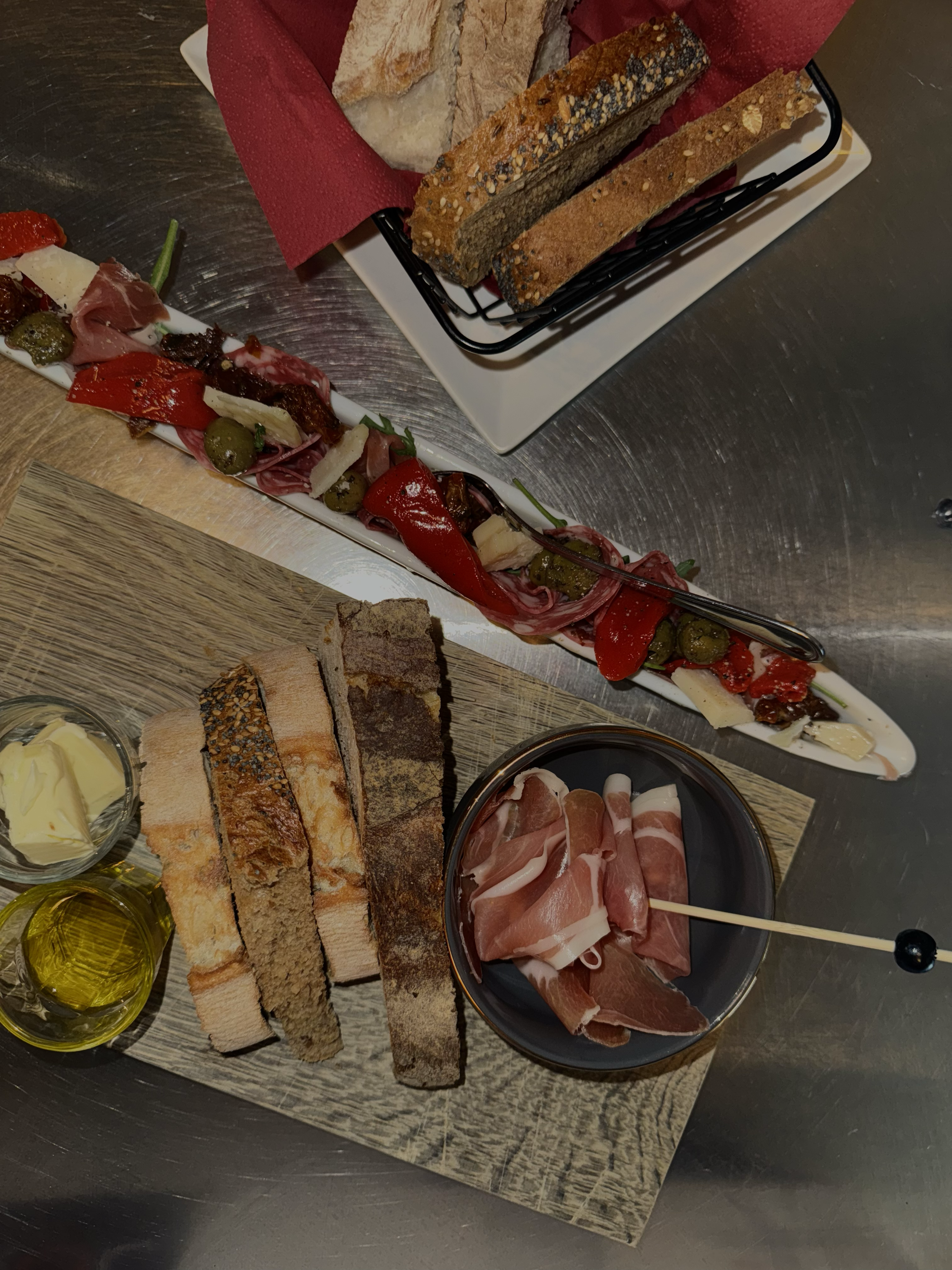 Brett mit verschiedenen Brotsorten, eine Schale mit in Scheiben geschnittenem Schinken, eine Schüssel mit Butter, zwei Flaschen mit Olivenöl, eine lange Platte mit Antipasti und Käse, eine kleine Schüssel mit Butter, eine Schüssel mit verschiedener r