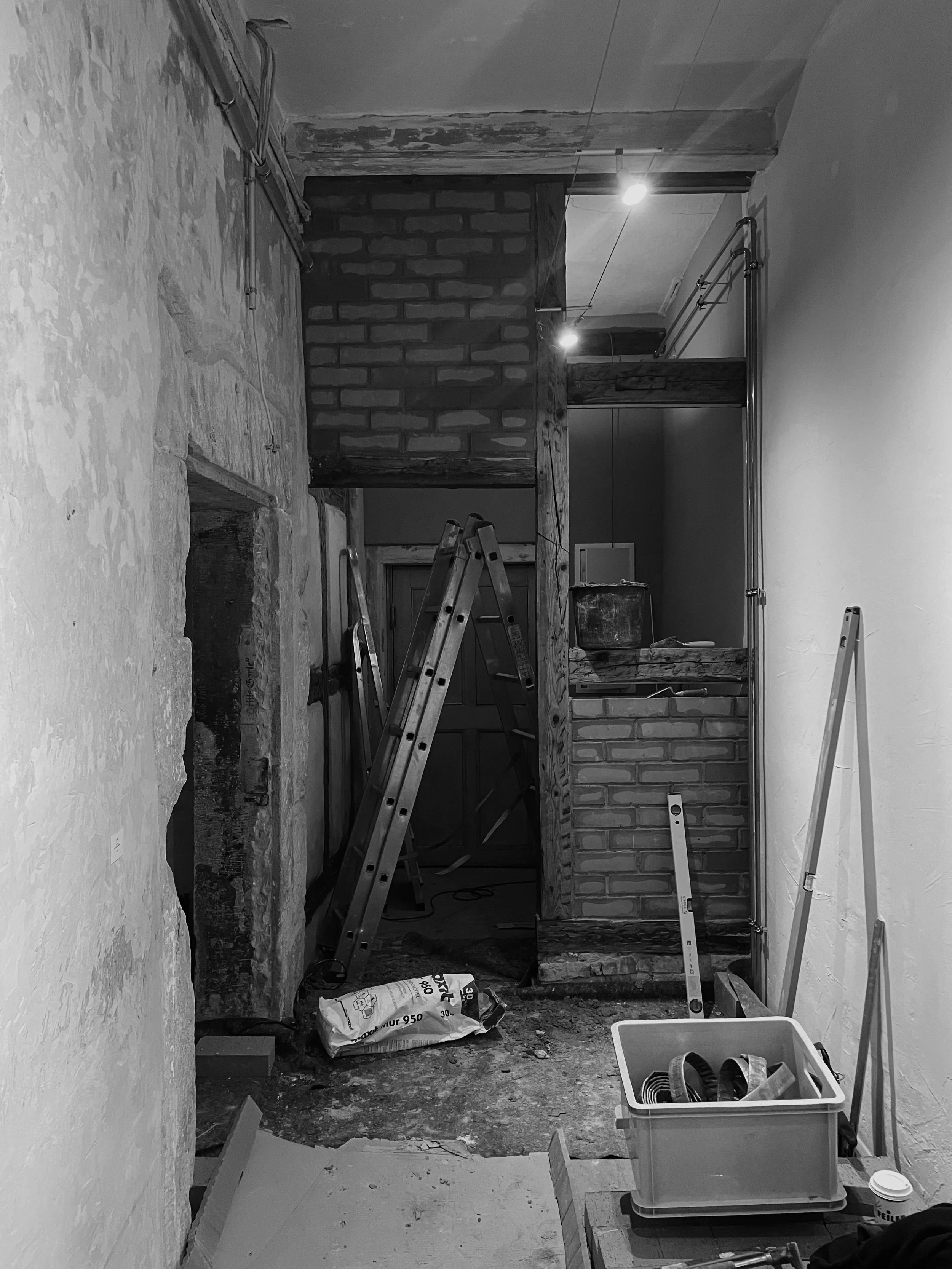 Innenraum einer Baustelle mit Ziegelwand, Leiter, Bauutensilien und Baumaterialien, Steckdosen und Rohre an der Wand, unsauberer Boden mit Bauabfällen.
