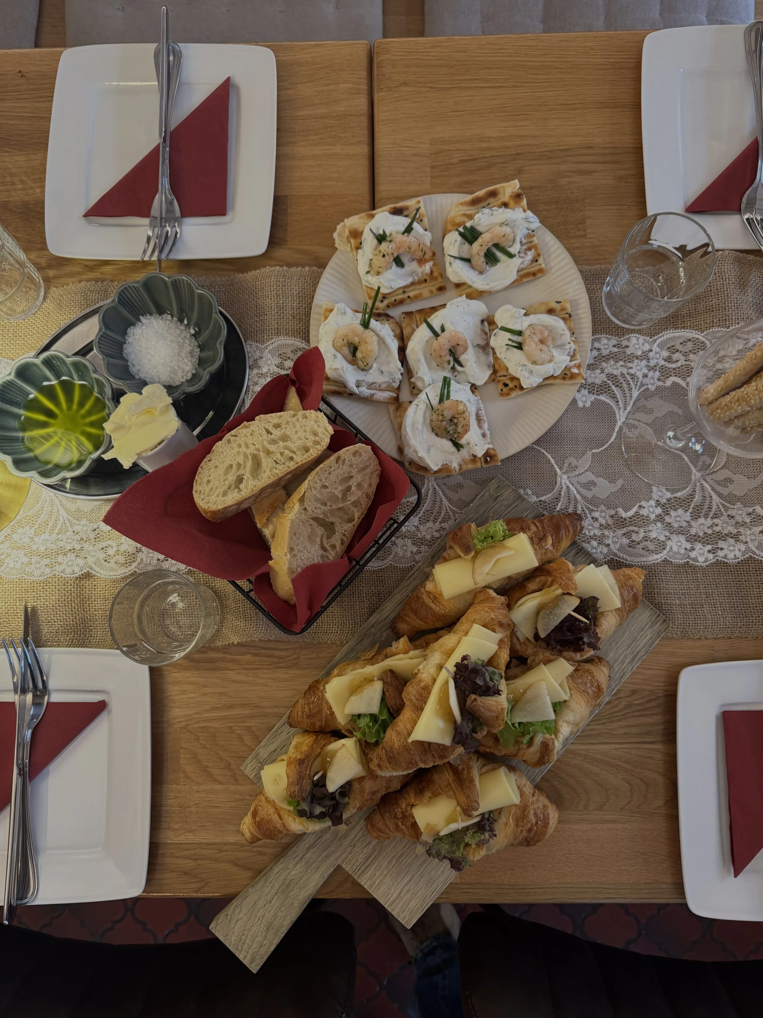 Ein Tisch mit verschiedenen Speisen: Ein Teller mit belegten Croissants, ein Teller mit Brot, Weingläser, ein Glas Wasser, Salz, Butter, eine Ecke mit Salz, servierte kleine Pizza, einen Korb mit Brot mit rotem Tuch, und Weißwein.