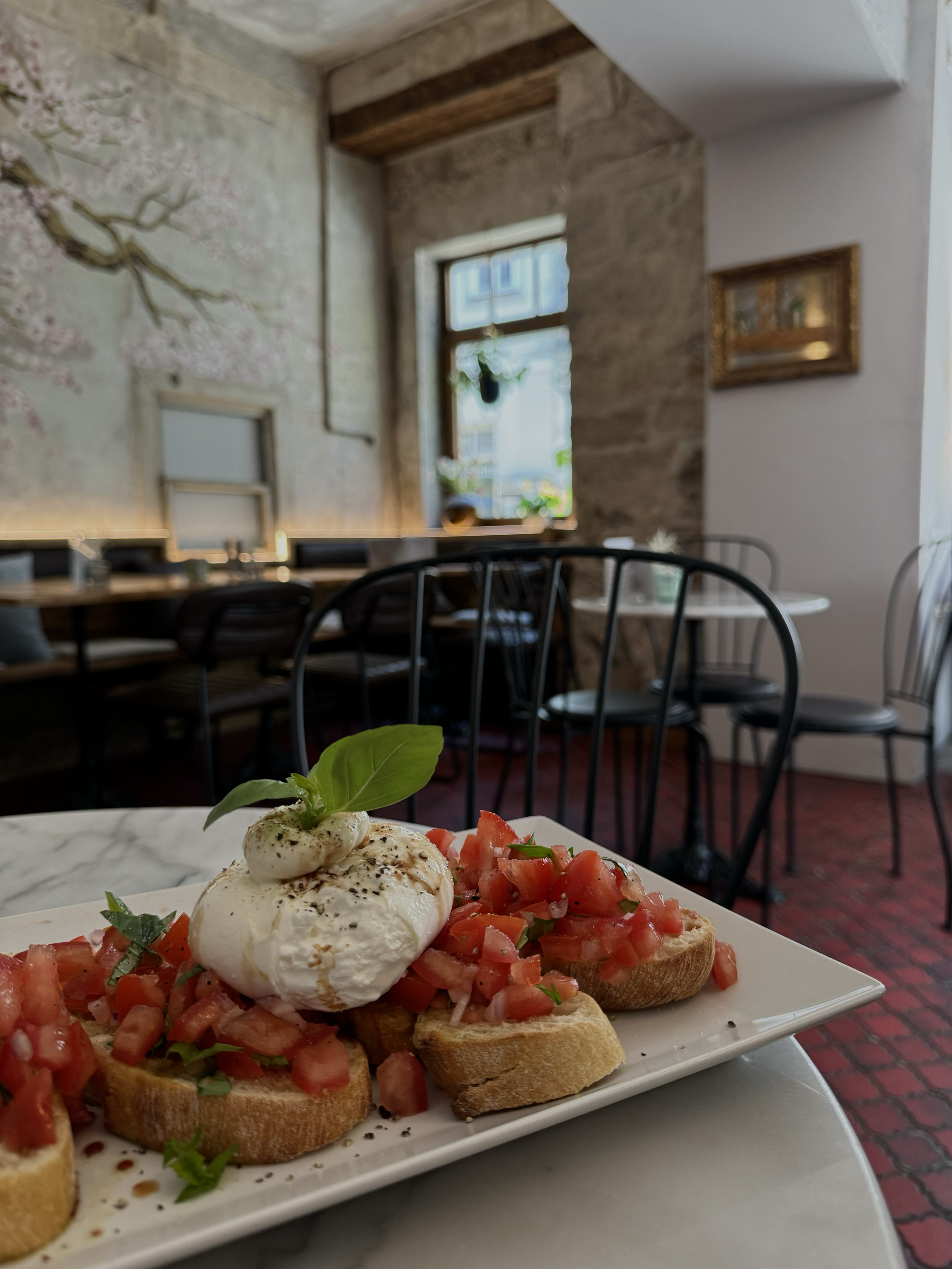Nahaufnahme von Bruschetta mit Tomaten, Basilikum und Mozzarella auf einem weißen Teller, im Restaurant. Im Hintergrund ein Fenster, Wand mit Blumenmuster, Tische und Stühle.
