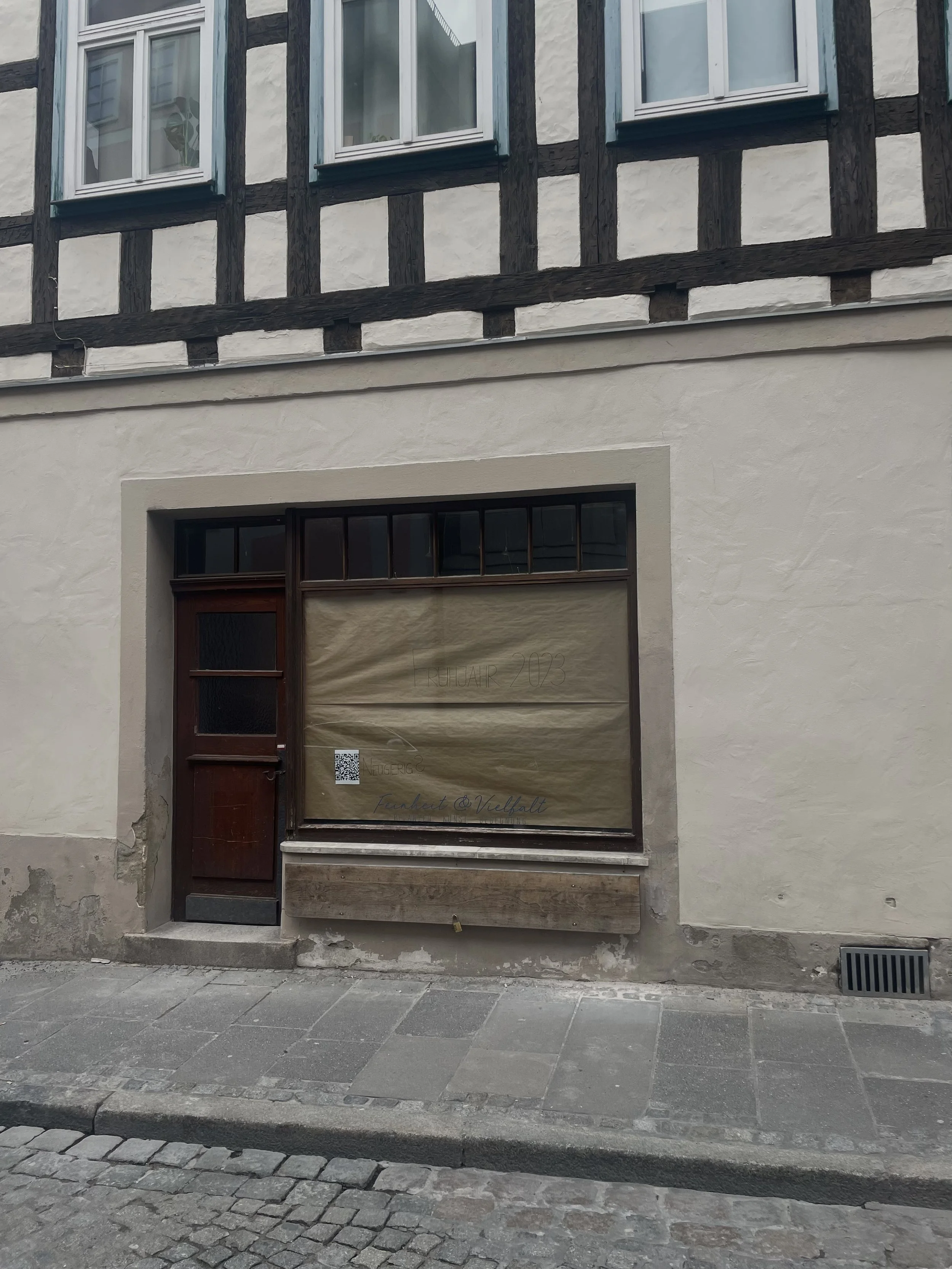 Ein altes Gebäude mit Holzrahmen und zwei Fenster im oberen Bereich. Das Erdgeschoss hat eine Tür und eine große Baustellenplane, auf der etwas geschrieben steht, sowie einen QR-Code. Der Gehweg ist gepflastert, und die Straße ist mit großen Steinen gepflastert.