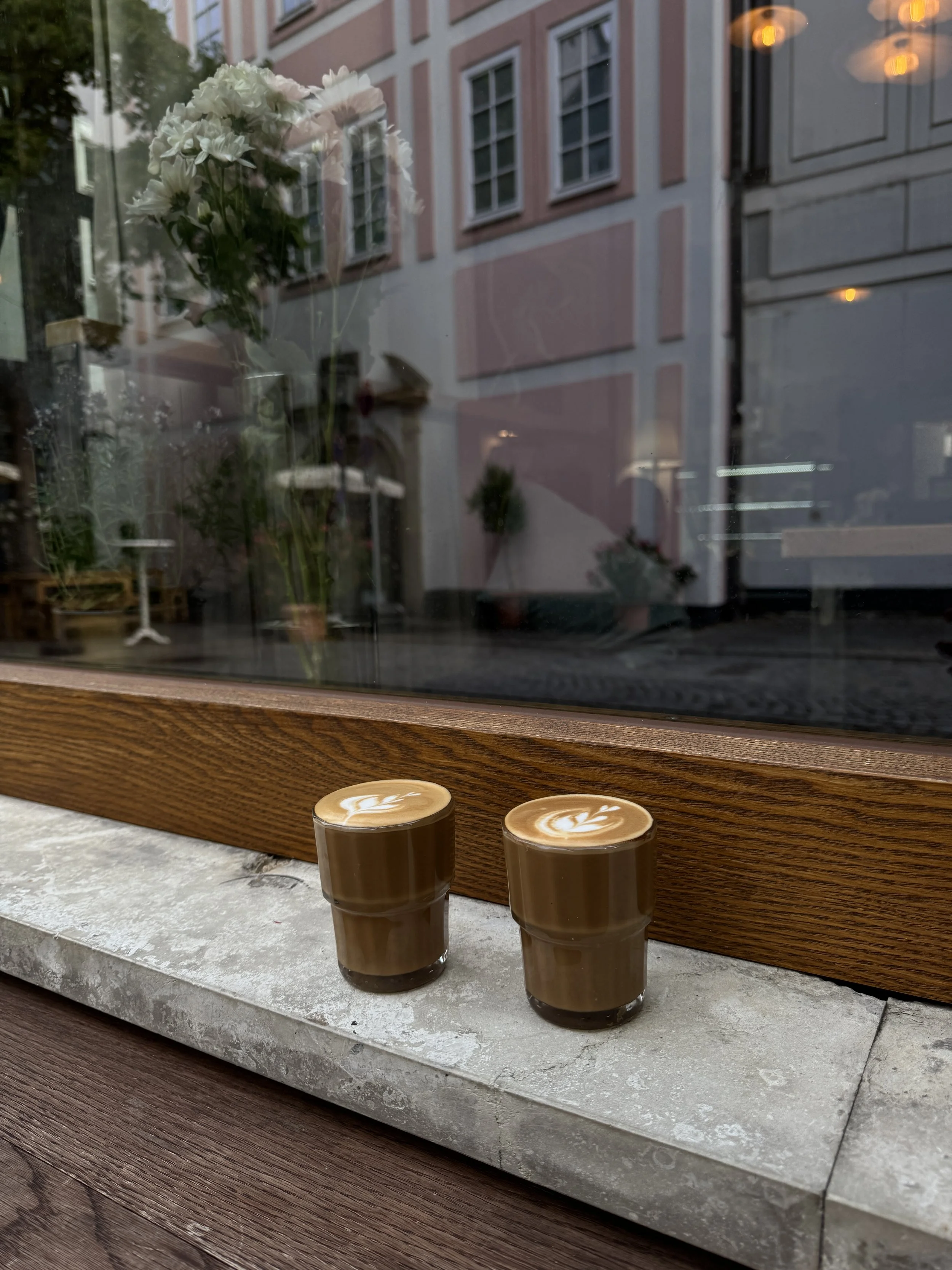 Zwei Tassen mit Kaffee mit Latte-Art auf einem Fenstersims, vor reflektierender städtischer Fassade und Blumen im Fenster