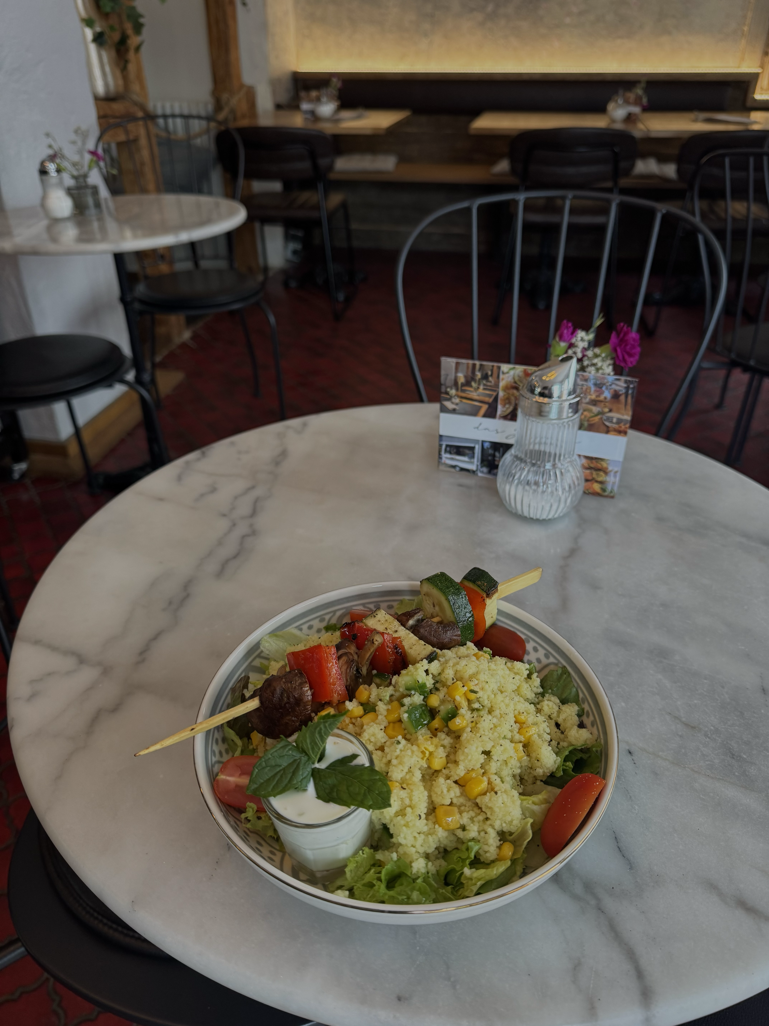 Ein Teller mit Couscous, gegrilltem Gemüse und Tomatenscheiben, serviert auf einem weißen Marmortisch in einem Restaurant.