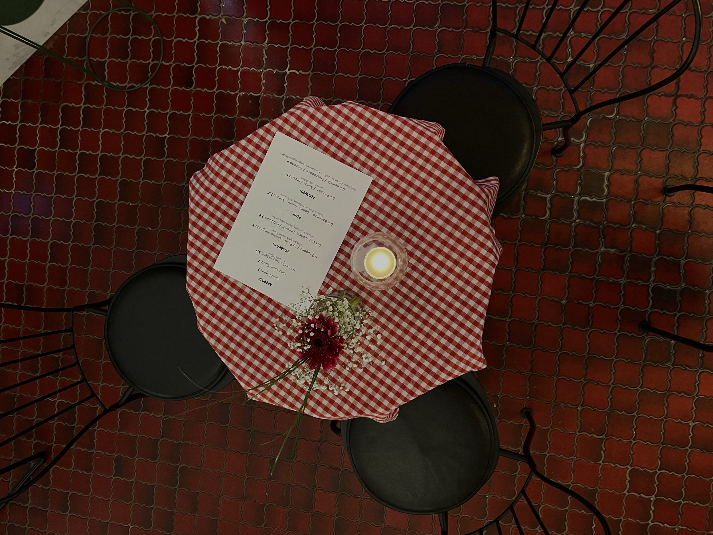 Ein Tisch mit rotem und weiß kariertem Tuch, eine Kerze, Blumen und eine Speisekarte. Um den Tisch herum schwarze Stühle, der Boden ist rot gemustert.