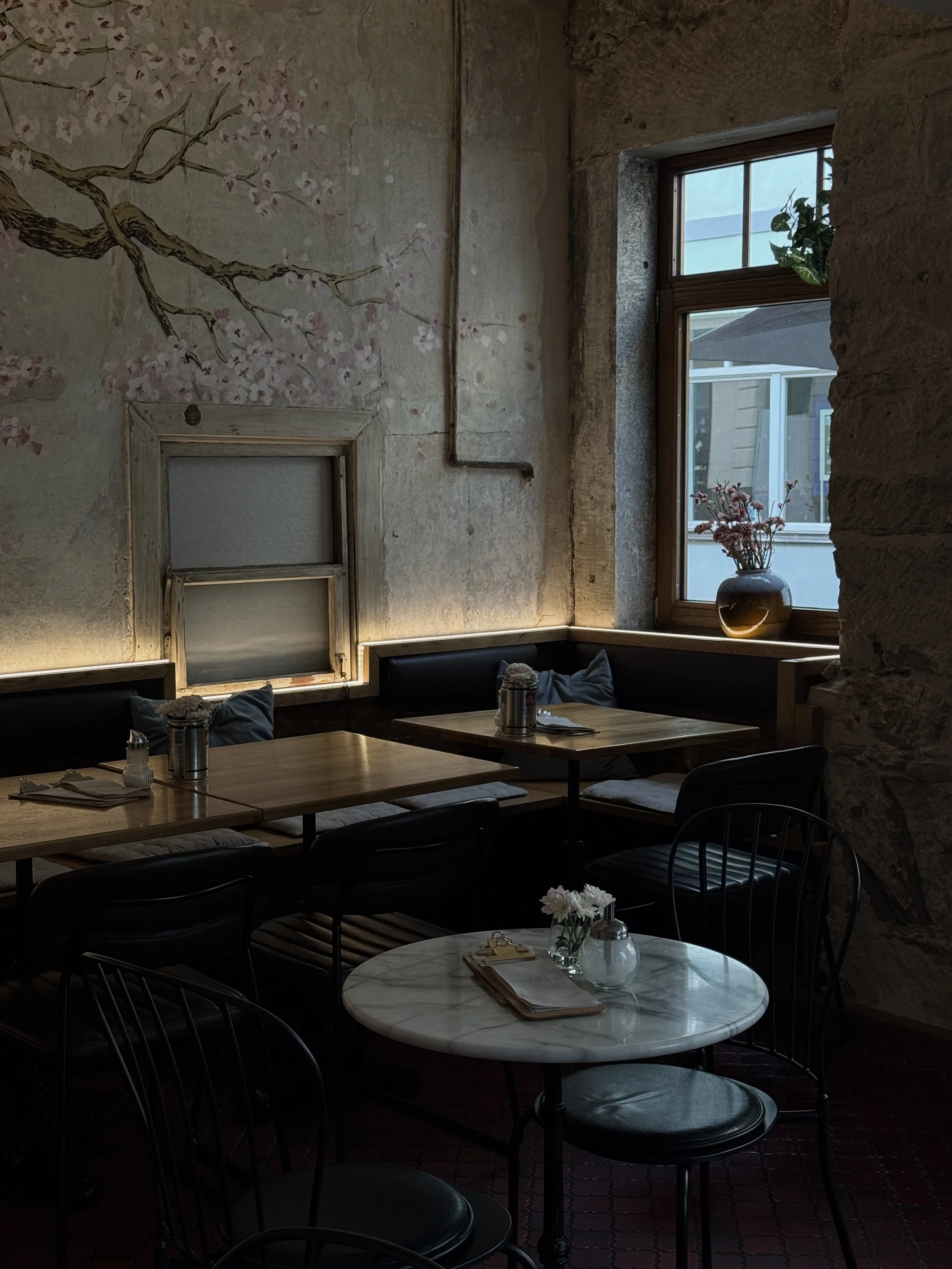 Ein gemütliches Café mit Holztischen, schwarzen Stühlen, einer weißen Marmortafel, einer Vase mit weißen Blumen, einem Fenster mit Sicht auf ein Haus, und einer Wanddekoration mit einem Baum mit blühenden Zweigen.