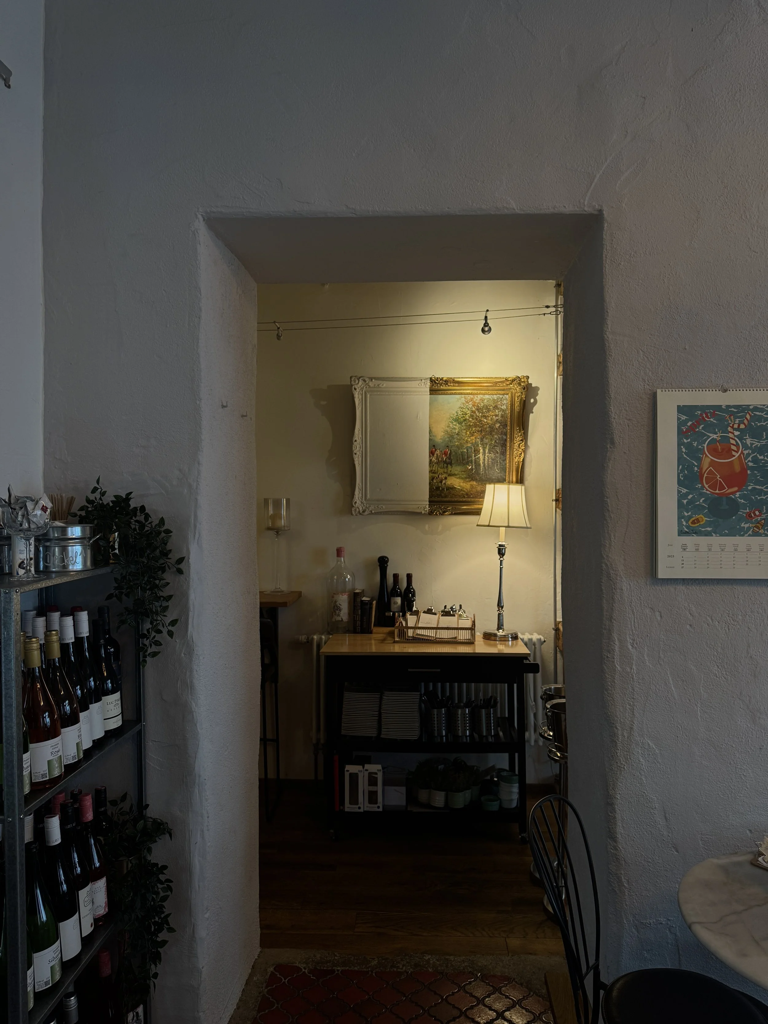 Einblick in ein gemütliches Café mit Weißwänden, Weinregal auf der linken Seite, einem Tisch und Stühlen im Vordergrund, einem Regal mit Weinflaschen, einem Gemälde an der Wand, einer Stehlampe, und einer beleuchteten Ecke im Hintergrund.