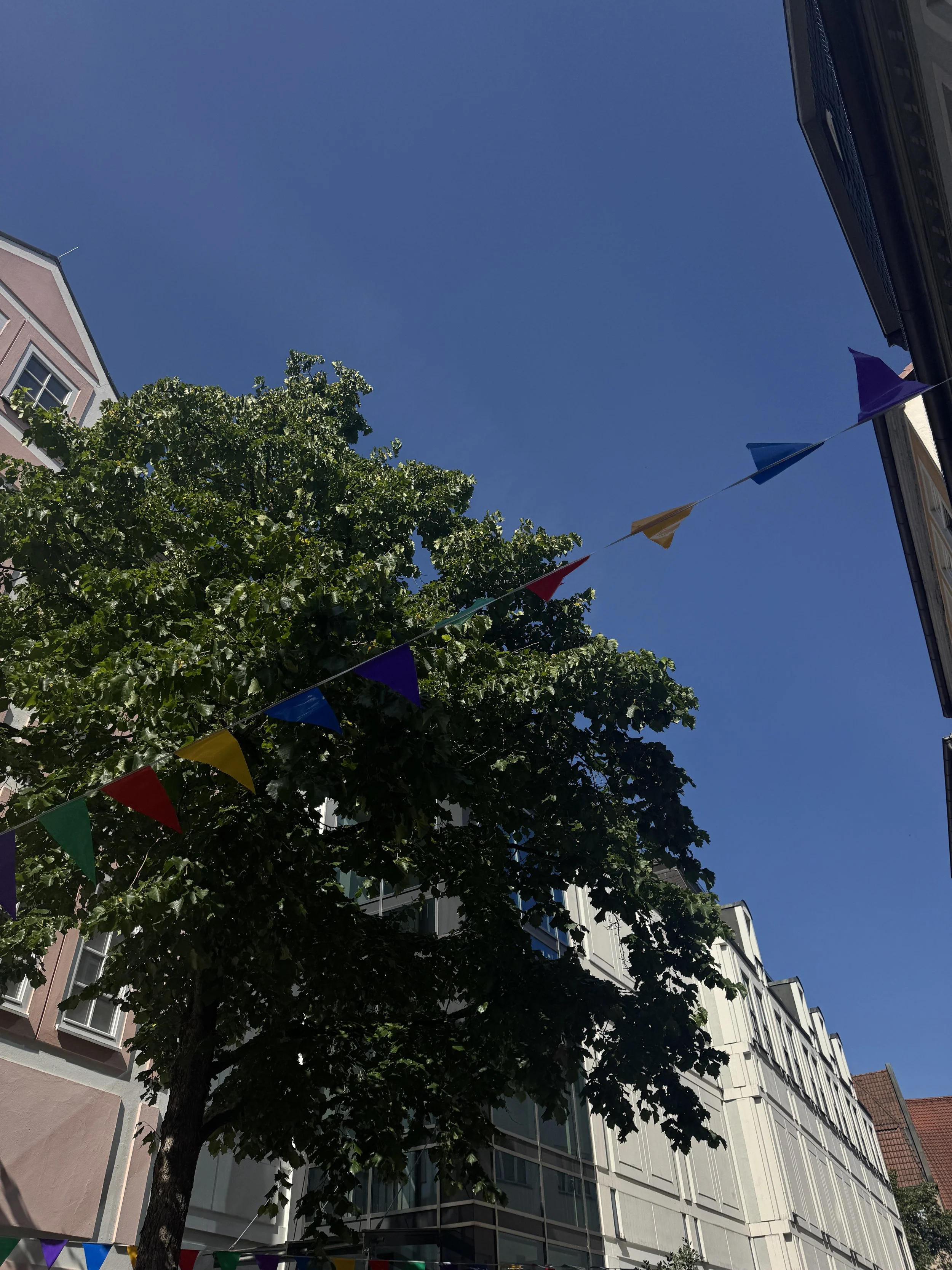 Blick nach oben auf einen klaren blauen Himmel, umgeben von Gebäuden und bunten Flaggen, die zwischen den Gebäuden aufgehängt sind.