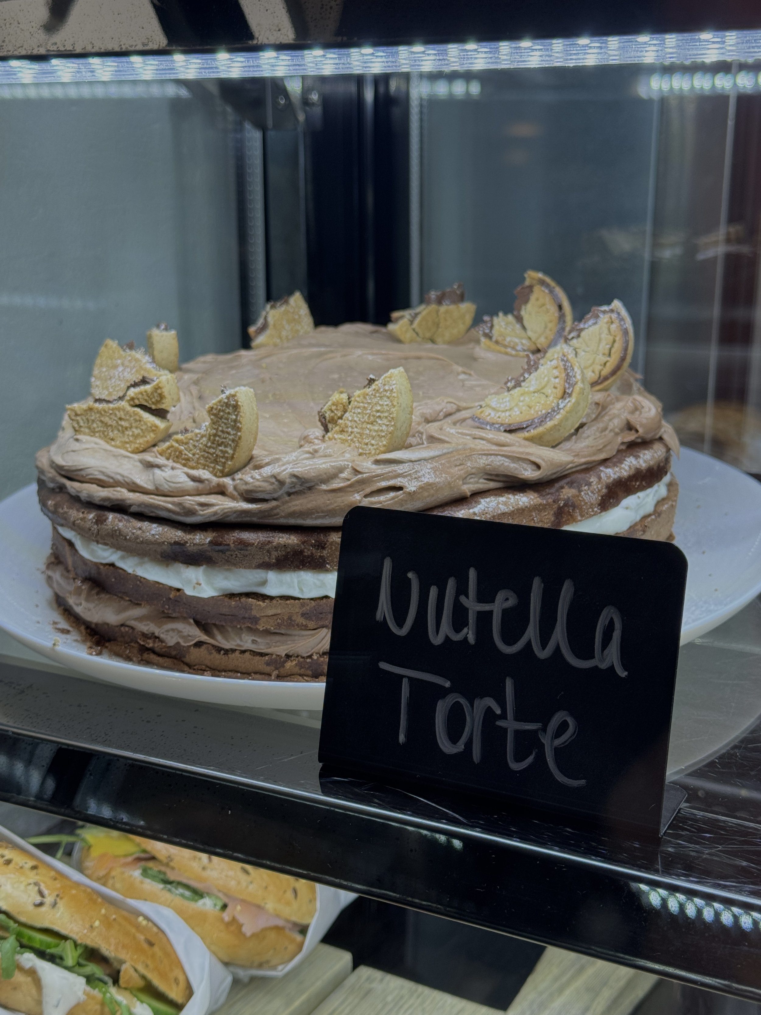 Ein Kuchen mit Schokoladenglasur und Biskuitboden, dekoriert mit Biskuitstücken und einer Füllung zwischen den Kuchenschichten. Es gibt einen kleinen schwarzen Zettel mit dem handgeschriebenen Wort 'Nutella'.