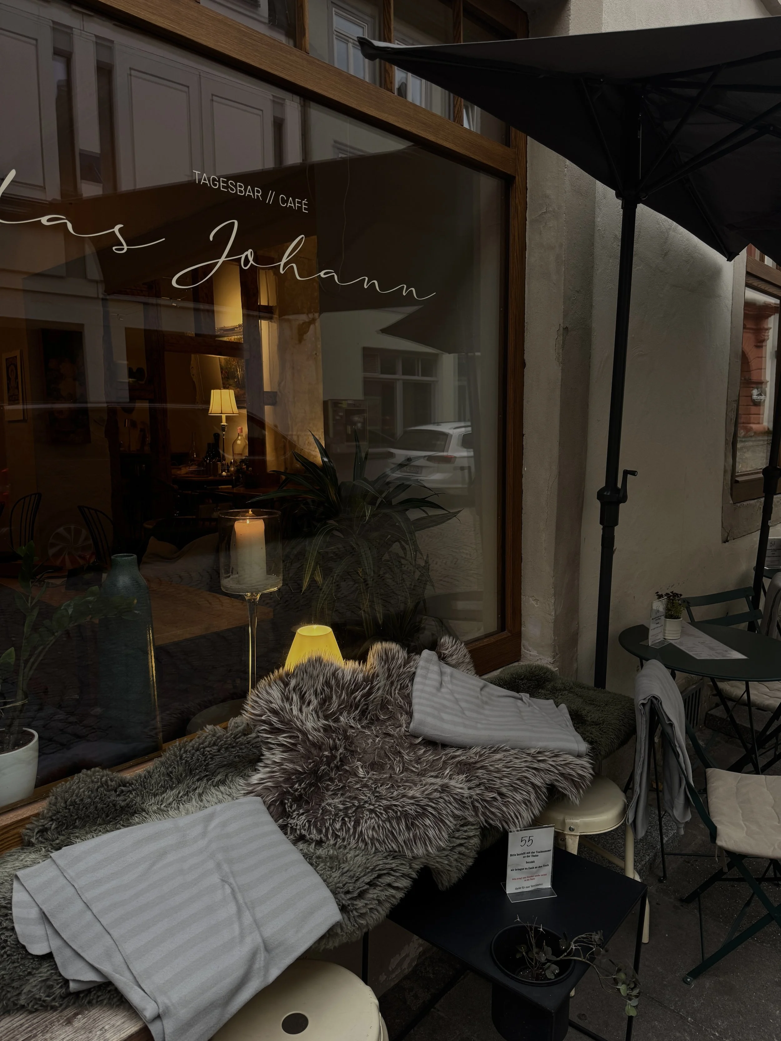 Straßencafé mit Blick auf ein Regal mit Decken, Tischtücher, eine Lampe, Pflanzen und andere Dekorationen im Inneren. Ein Außenbereich mit Stühlen, Tisch und Sonnenschirm, Blick durch die Fenster auf eine Straße mit Autos.