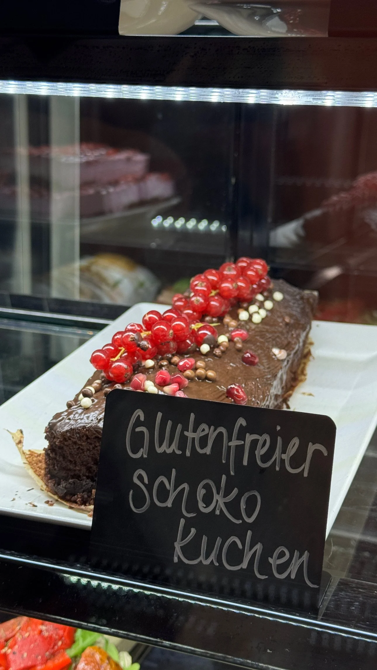 Schokoladenkuchen mit roten Johannisbeeren und weißen Schokotropfen in einer Vitrine.