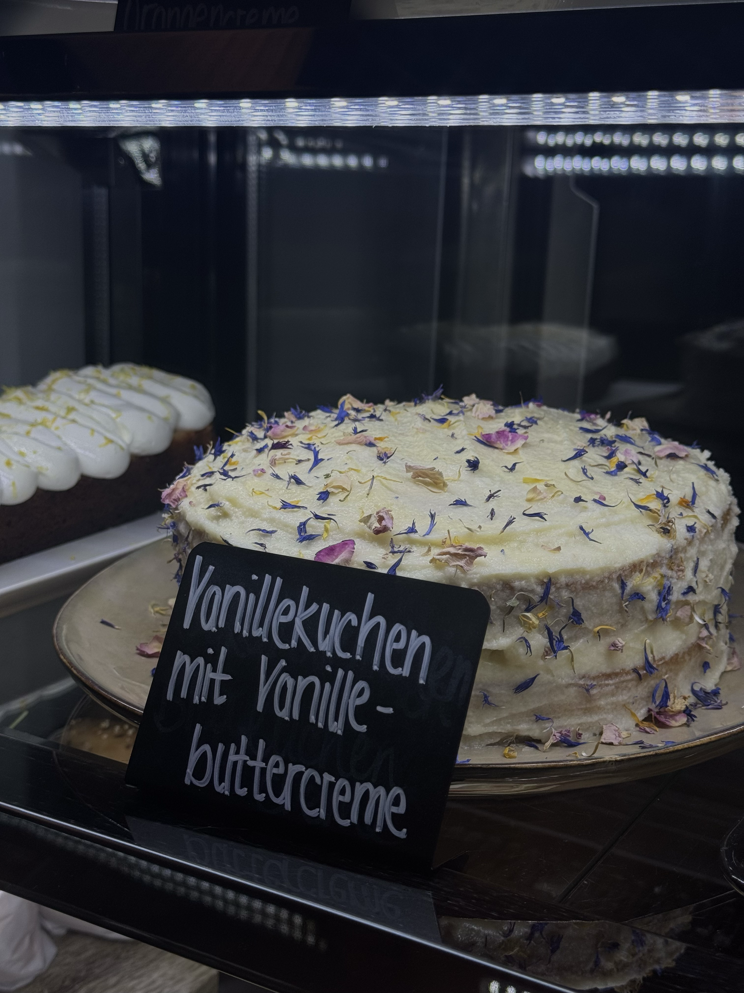 Ein Kuchen mit weißen Frosting und Blütenblättern. Daneben eine Platte mit weiteren Keksen oder Kuchenstücken. Ein schwarzes Schild mit weißer Schreibschrift, auf dem steht: "Vanillekuchen mit Vanille-"