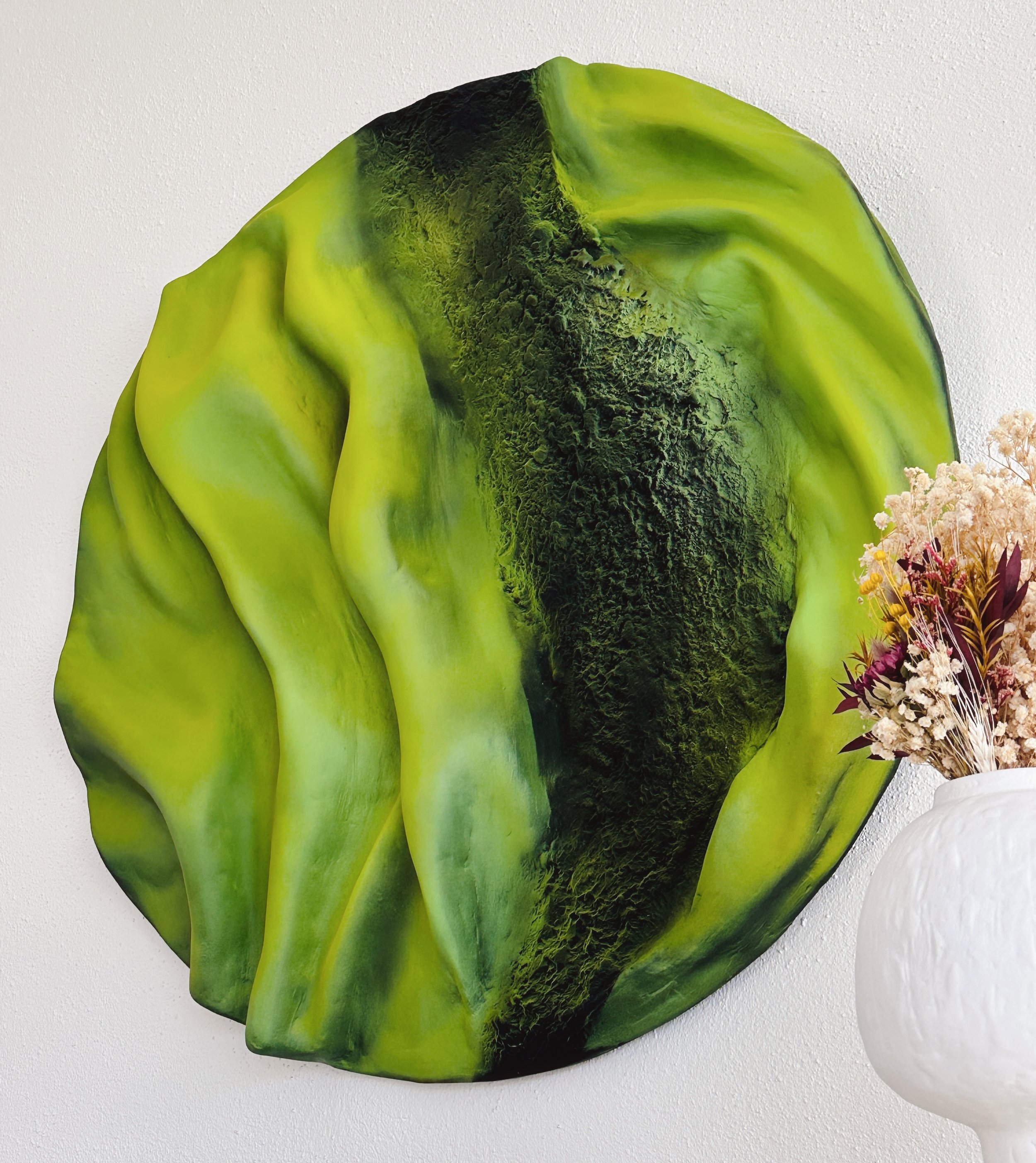 Close-up of a large, vibrant green leaf with textured dark green area in the center, against a plain white wall background. Part of a small white vase with pink and yellow flowers is visible on the right side.