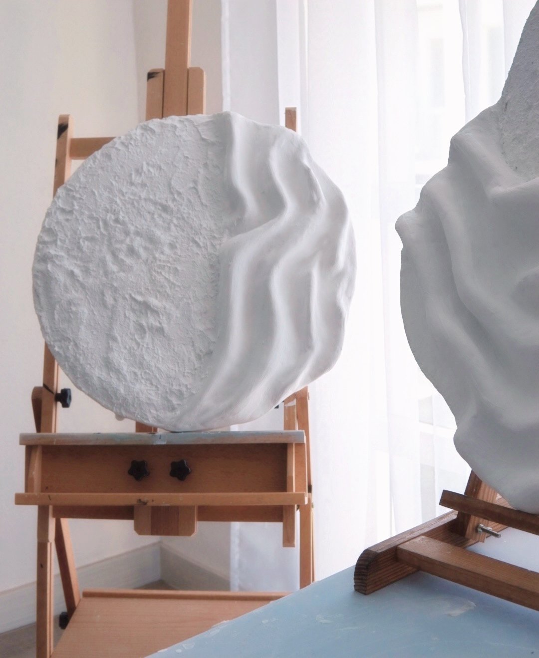 Close-up of a white plaster sculpture of a face with detailed features, placed on a wooden easel in a well-lit room with sheer curtains.