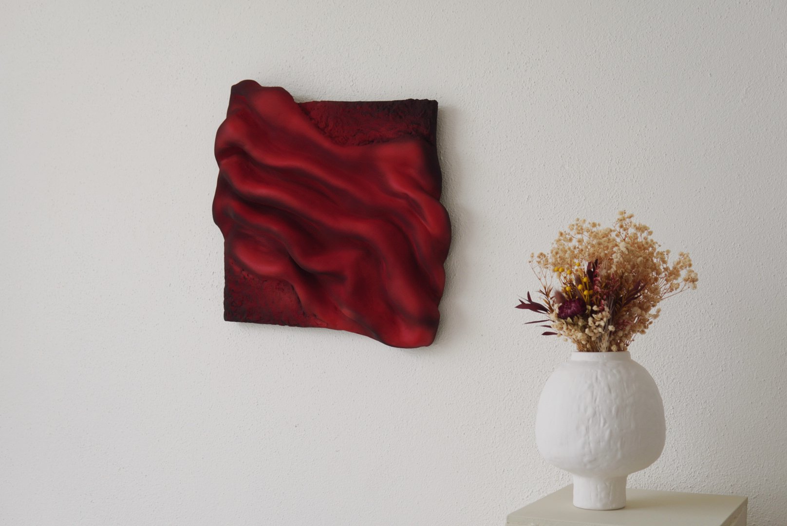 A white textured ceramic vase filled with dried flowers, placed on a neutral-colored table against a white wall. A modern red and black abstract wall art hangs on the wall.