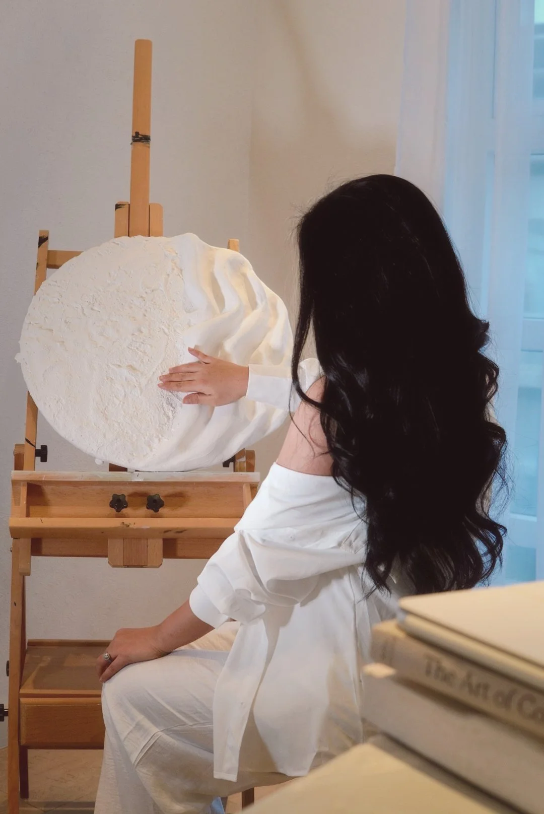 A woman with long black wavy hair in a white dress sitting in front of an easel, touching a large white textured circular art piece on display.