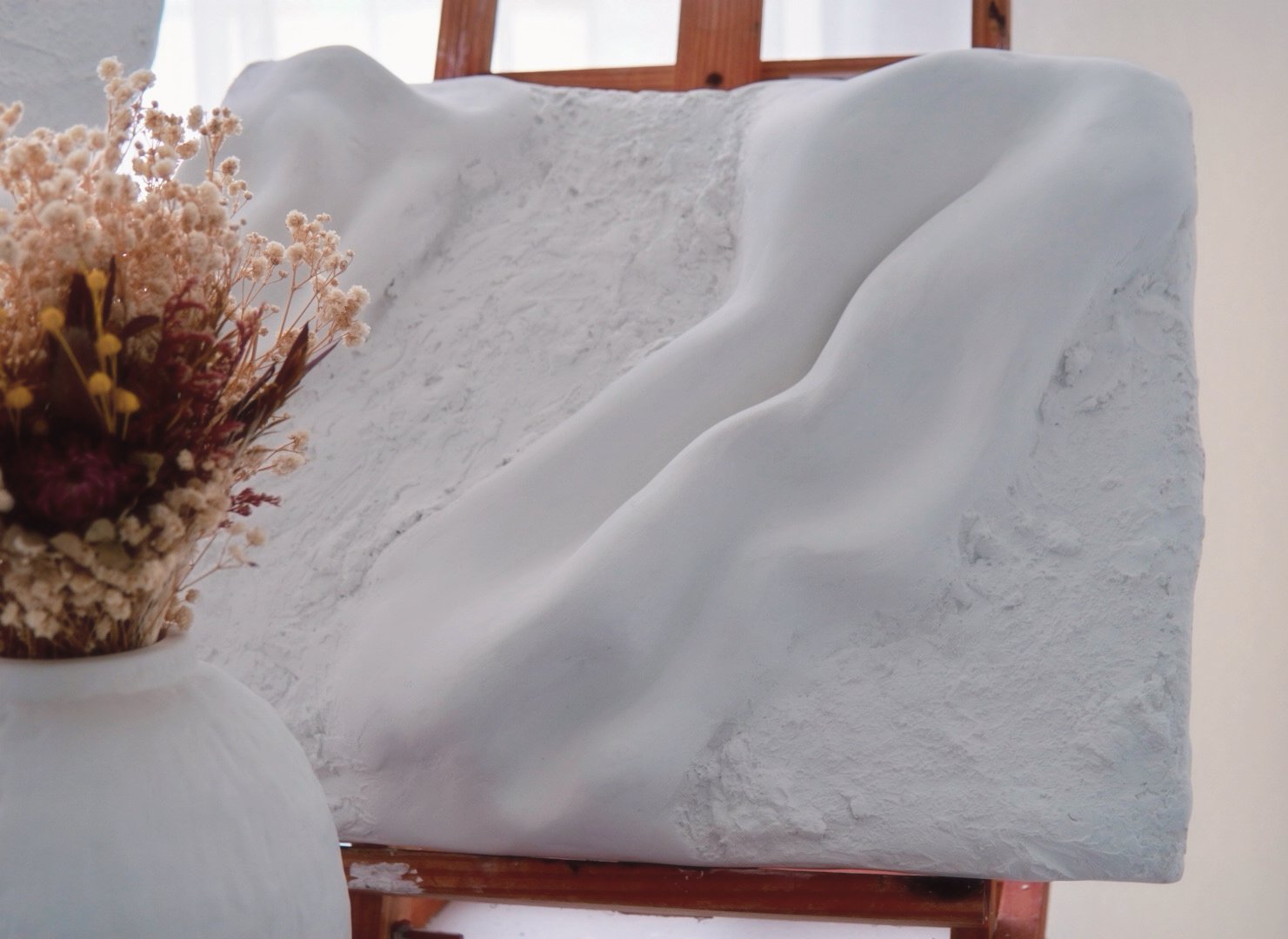 A white sculpture of a mountain or rock formation with a textured surface, placed on a wooden stand. A white vase with dried flowers is partially visible in the foreground.