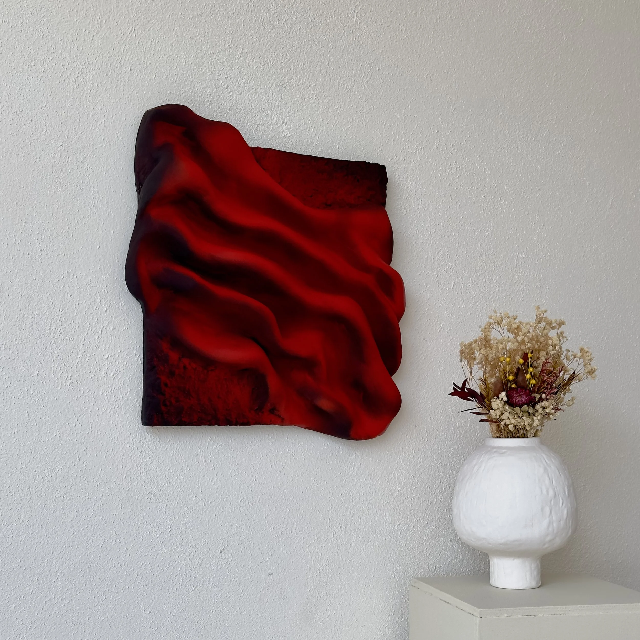 A red artificial flower with black streaks in a white vase placed on a shelf against a white textured wall.