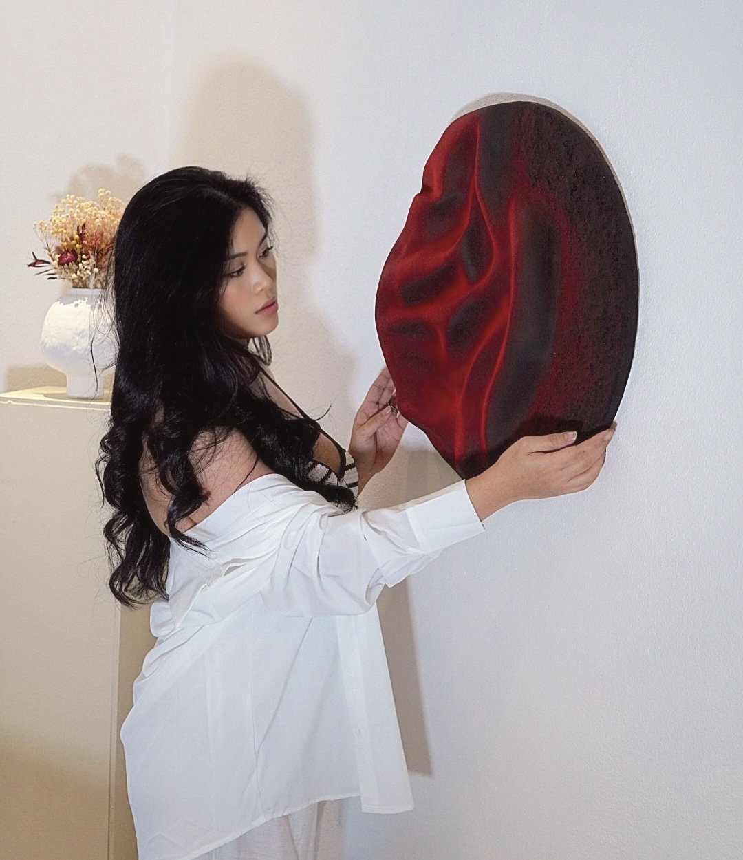 A woman with long black hair wearing a white shirt is standing and looking at a large, abstract red and black art piece on the wall.