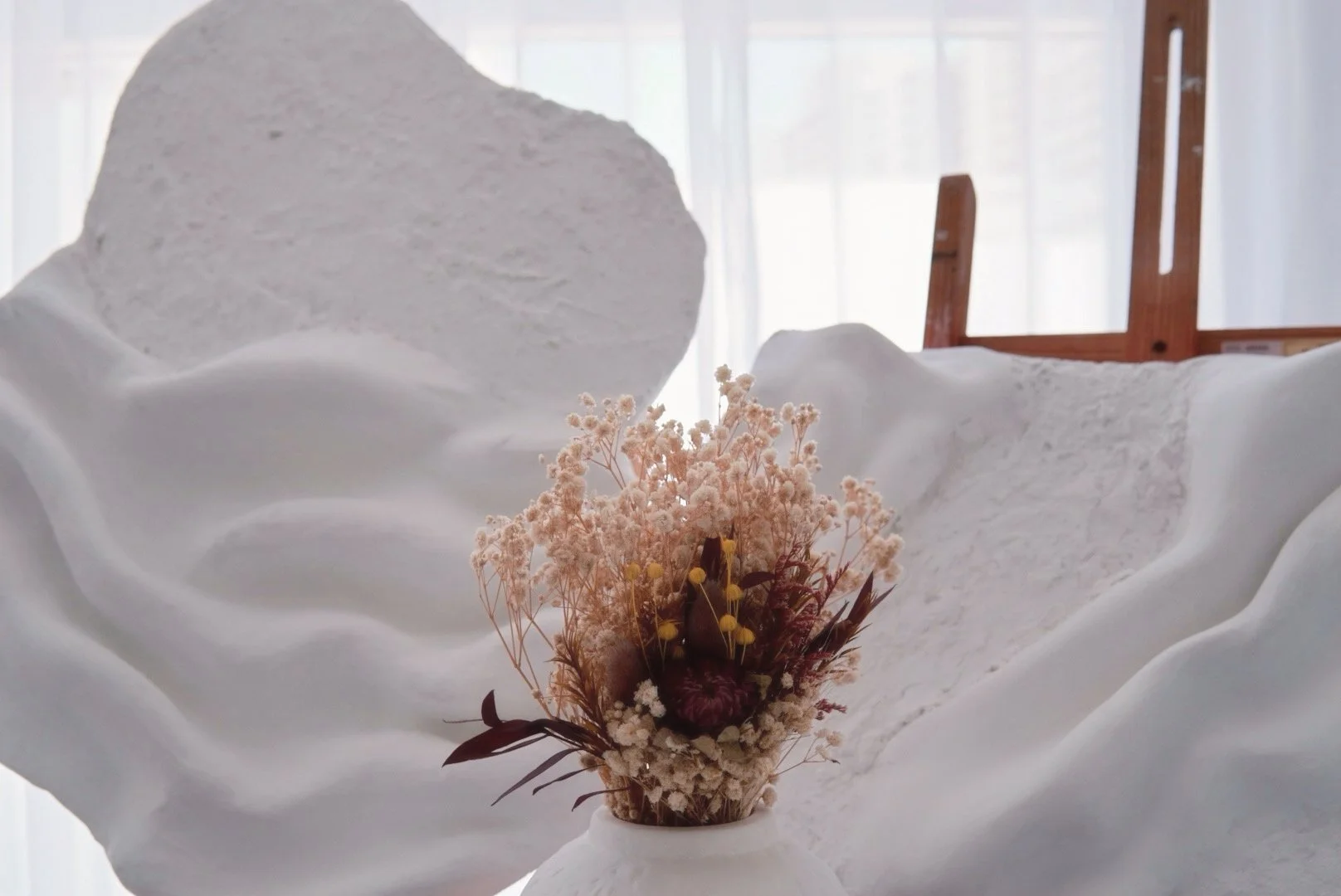 A white textured sculpture resembling a reclining figure with a bouquet of dried flowers in a white vase in front of it, set against a softly lit background with a wooden easel to the right.