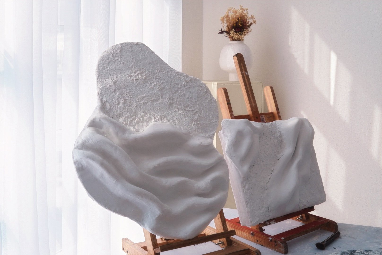 Two white sculpted busts with smooth, textured surfaces on wooden easels in a bright room with sheer curtains and a vase of dried flowers in the background.
