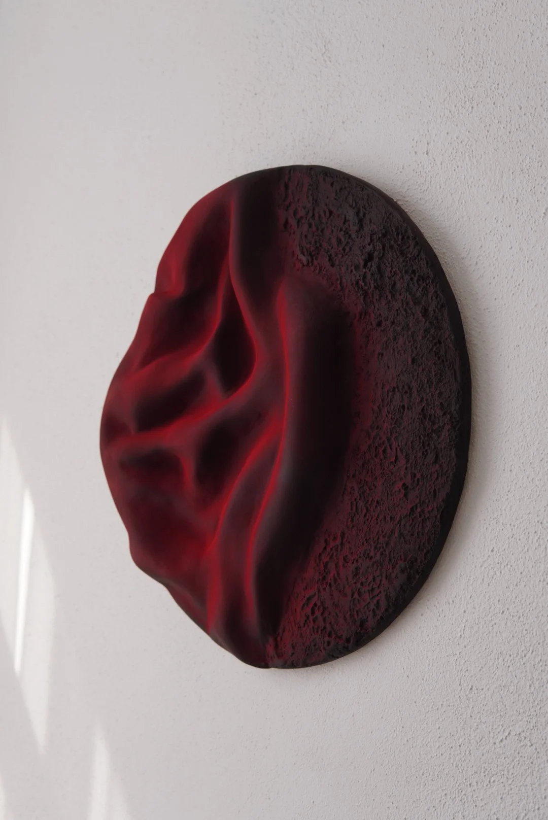 A circular wall sculpture resembling a human ear with black and red textured and smooth surfaces, mounted on a white wall.