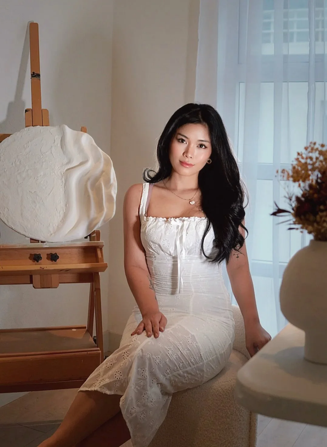 A woman with long black hair wearing a white dress sitting on a beige sofa in a well-lit room, with an art easel and sculpture on either side.