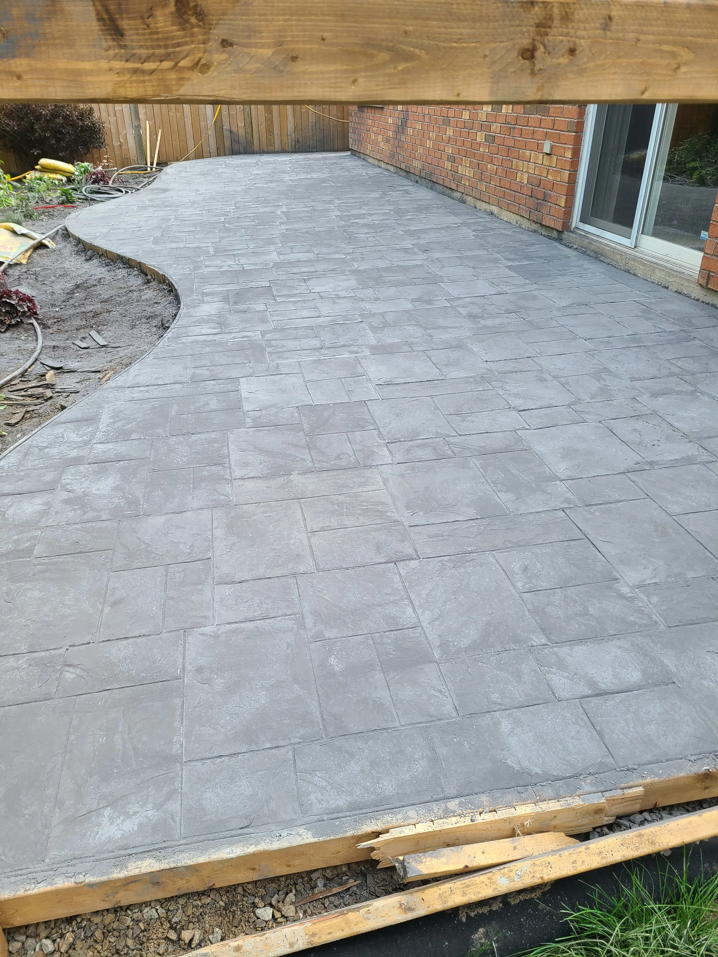 A newly constructed concrete patio with textured paving stones, adjacent to a brick house with a sliding glass door, surrounded by a wooden fence, construction materials, and tools.