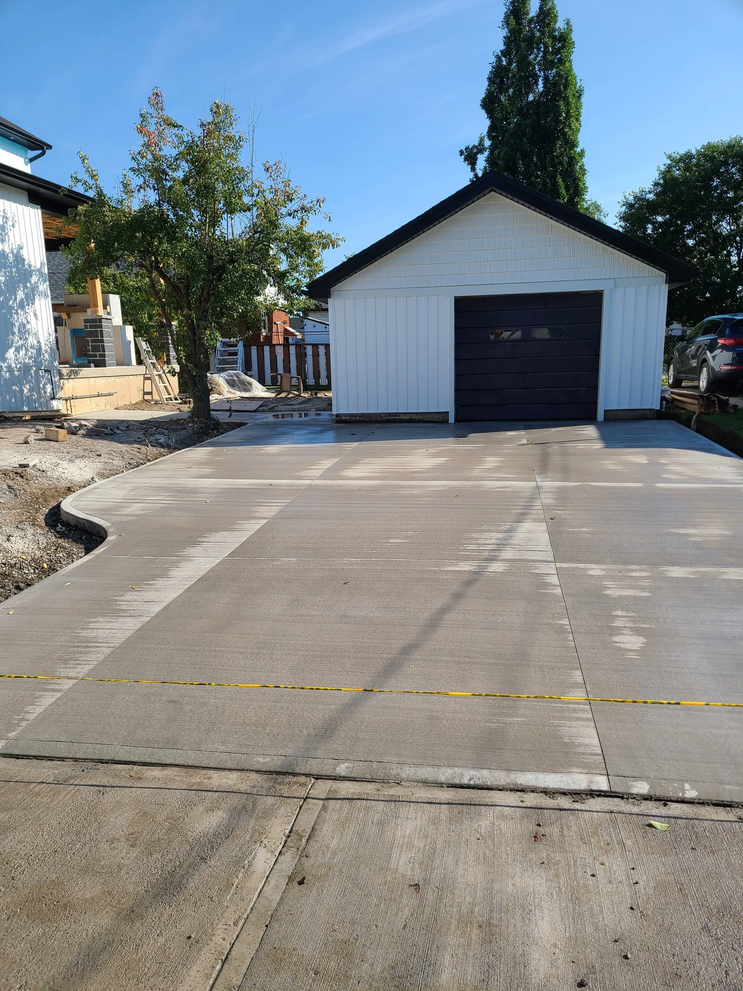A small white garage with a black door, on a paved driveway, with trees and a house under construction in the background, on a clear sunny day.