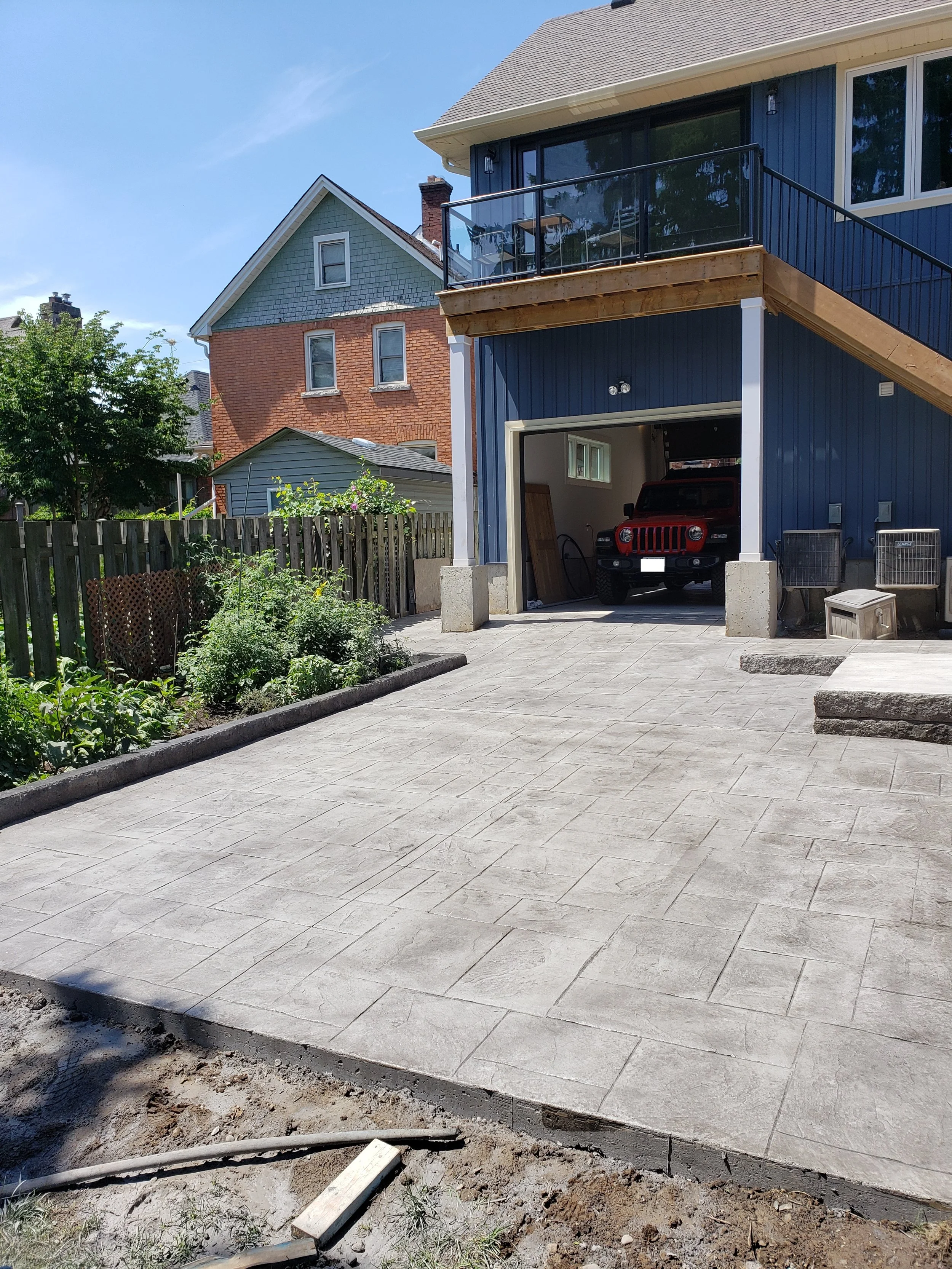 A newly paved concrete driveway in front of a house with construction work ongoing, with grass and plants on the left and a blue house with a balcony on the right.