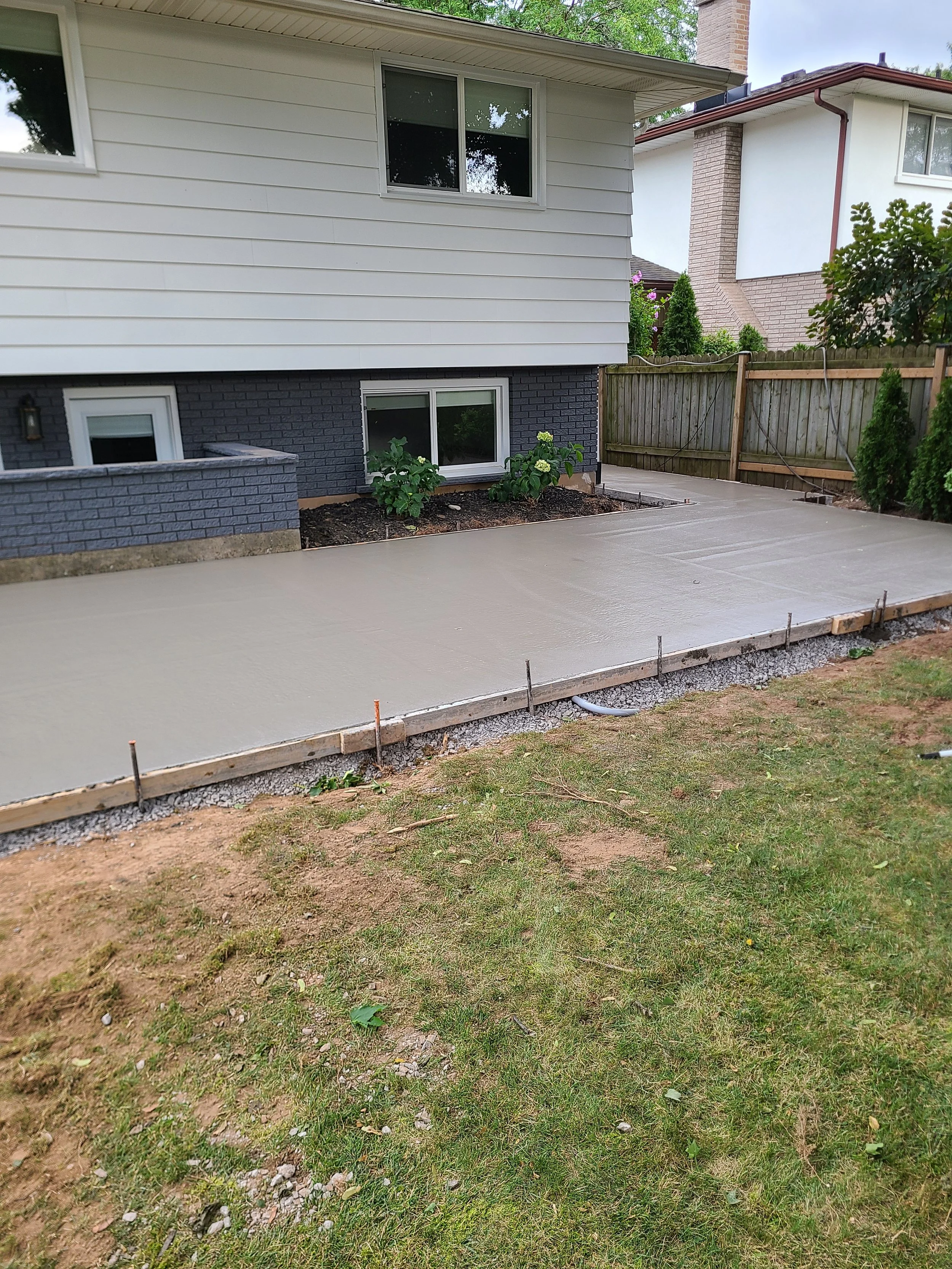 Freshly poured concrete patio in front of a house, with some plants along the house and a grassy yard adjacent.