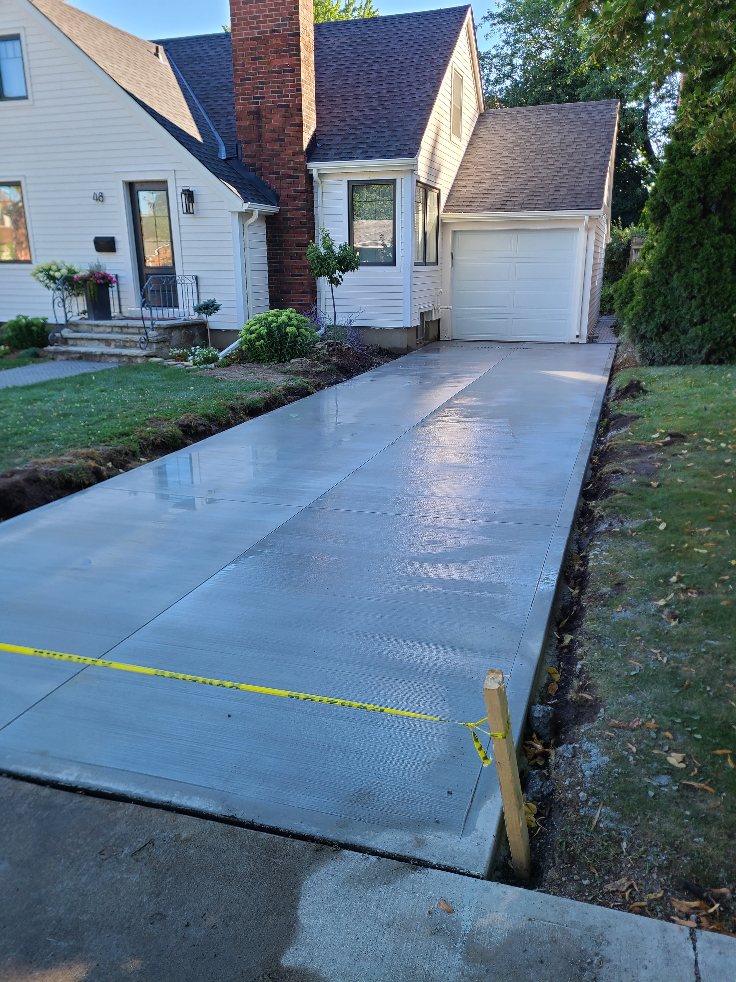 Freshly poured concrete driveway with a yellow safety tape border, next to a house with white siding and a brick chimney, on a sunny day.