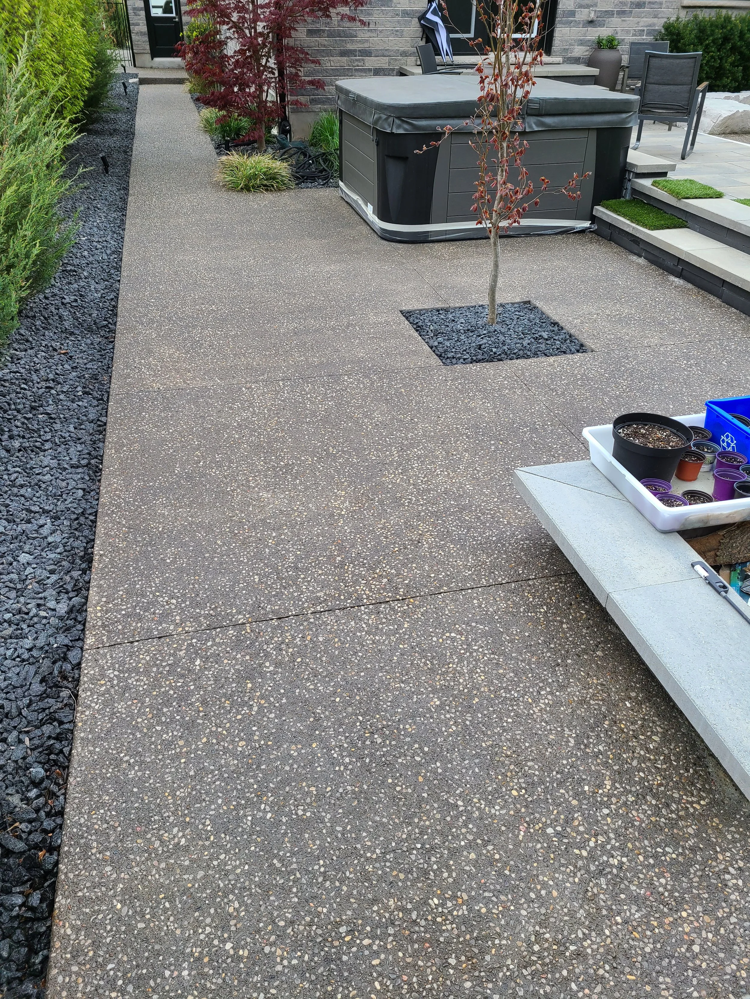 A backyard patio with a textured concrete floor, small plants, and a hot tub near the steps. There are chairs, a potted plant, and gardening supplies visible.
