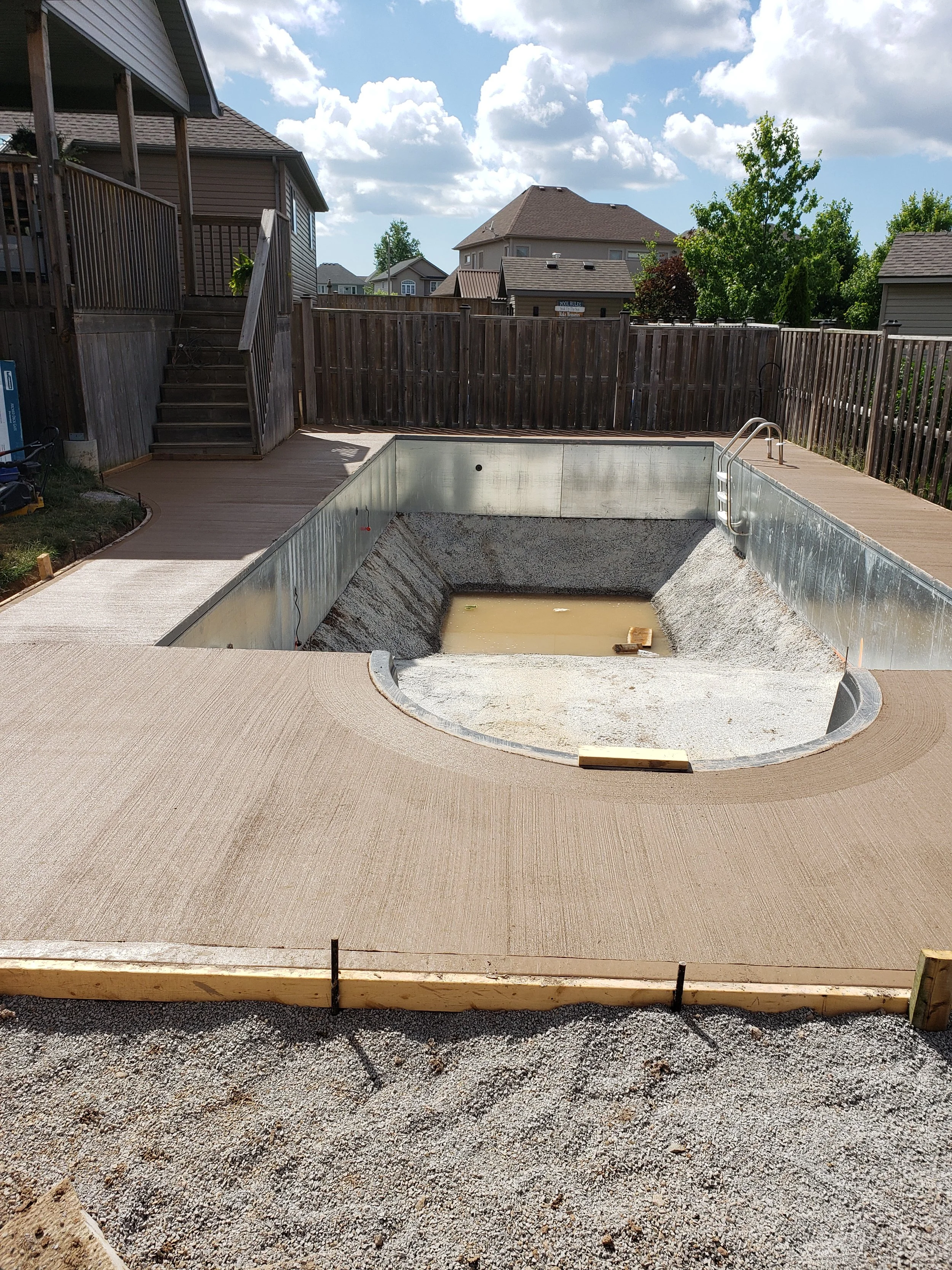 A backyard swimming pool under construction with an incomplete concrete deck, surrounded by a wooden fence and residential houses in the background.
