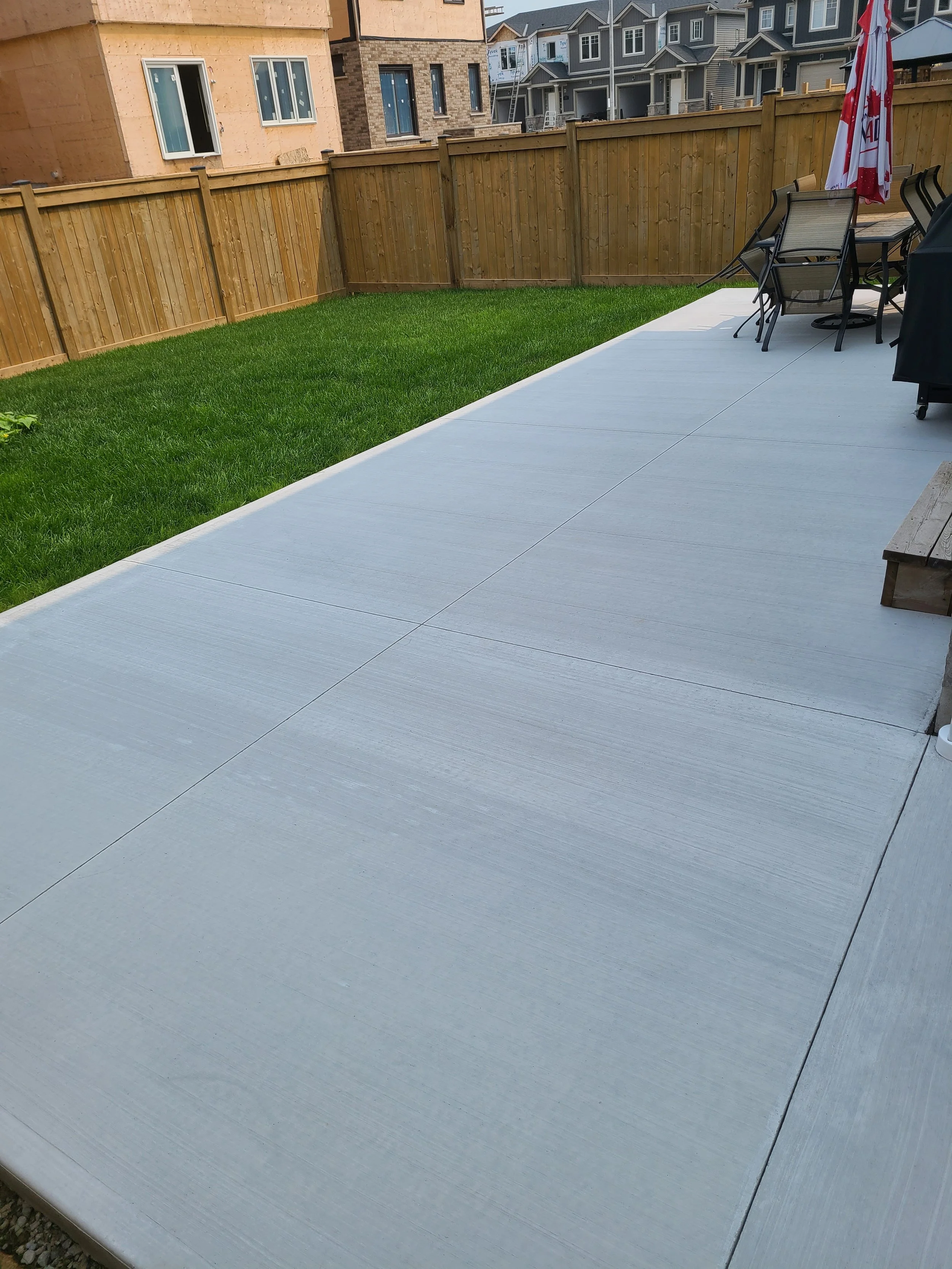 Backyard with a concrete patio, green grass, a wooden fence, outdoor table with chairs, and multicolor houses in the background