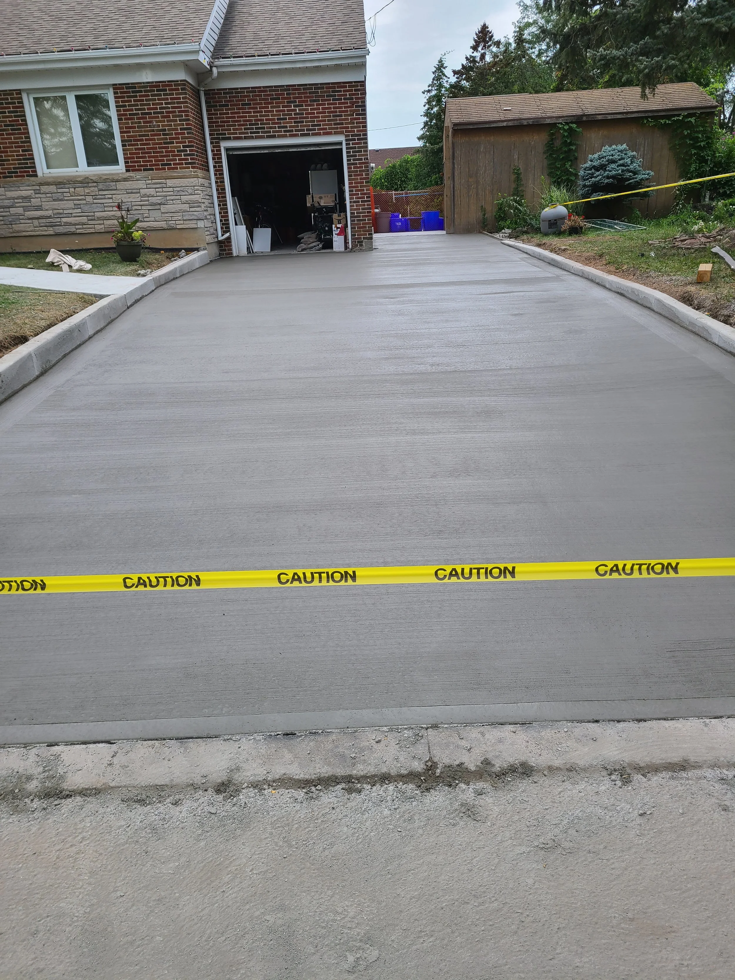 Freshly poured concrete driveway with yellow caution tape at the edge, in front of a house with a brick facade and a garage door open to storage.