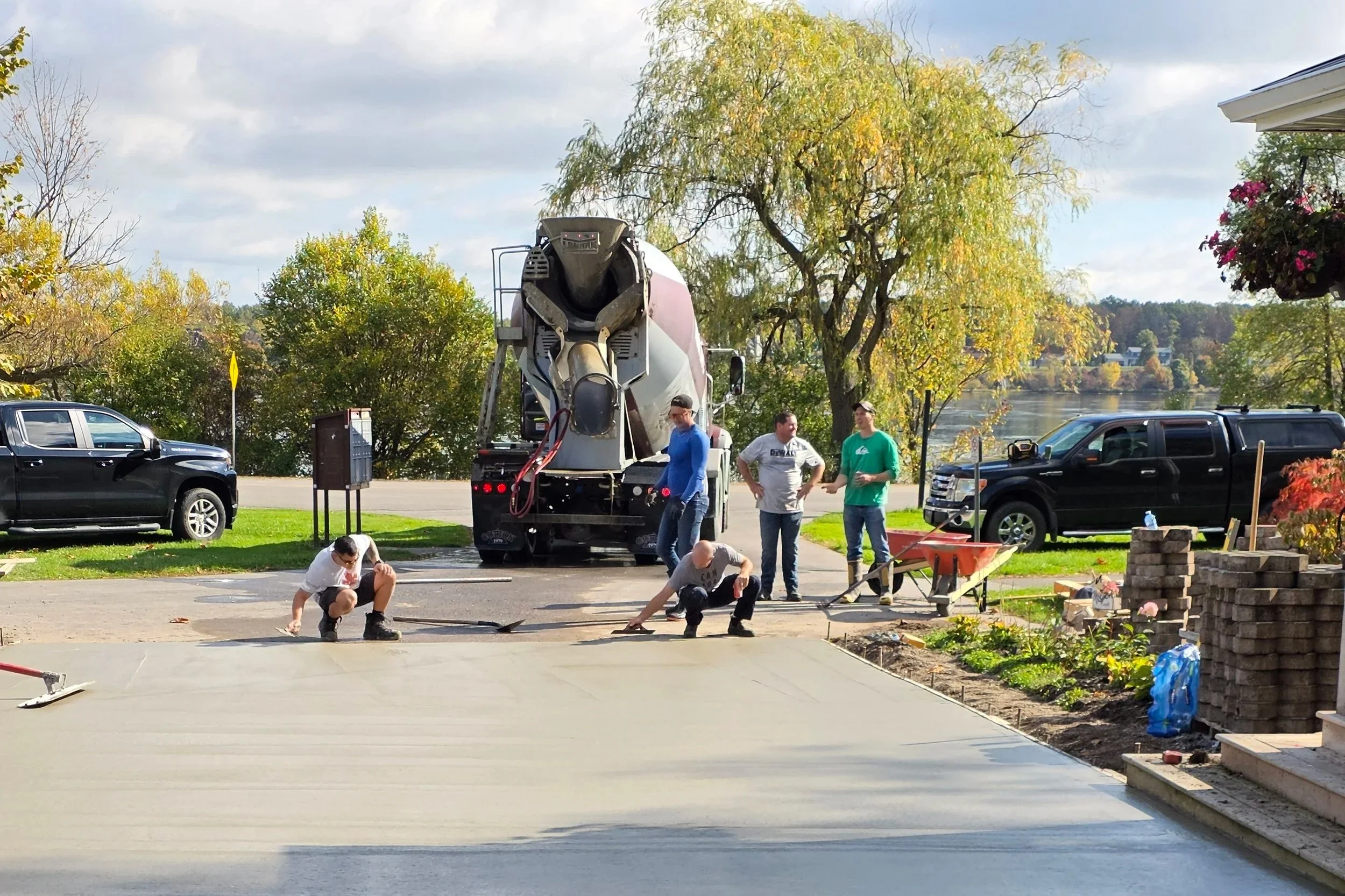 People working on a concrete driveway in a residential area with trees, parked cars, and a river in the background.