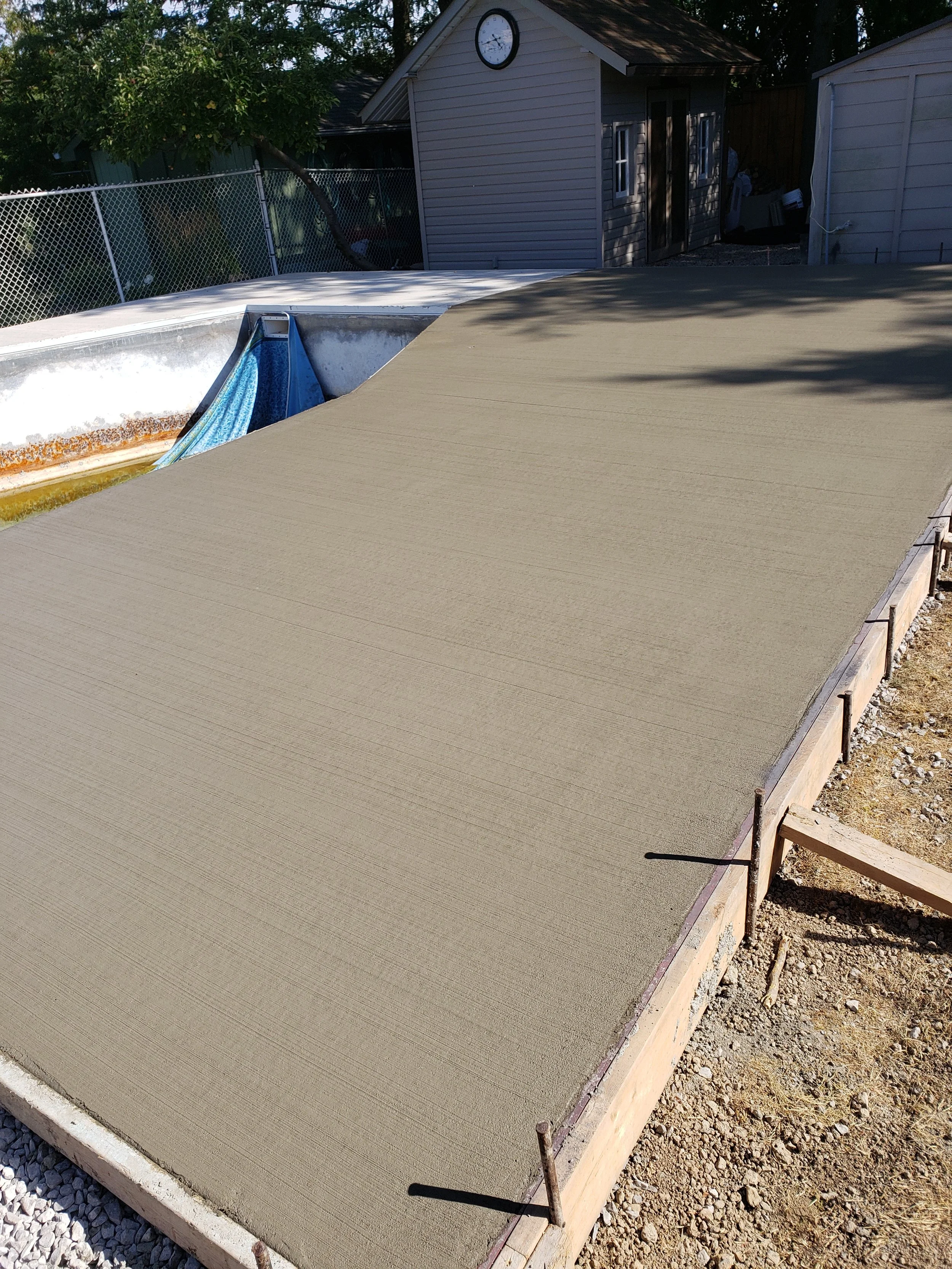 A swimming pool with a newly poured concrete deck, surrounded by a wooden frame and gravel on one side. In the background, there is a small house with a clock on the wall, a chain-link fence, trees, and some outdoor equipment.