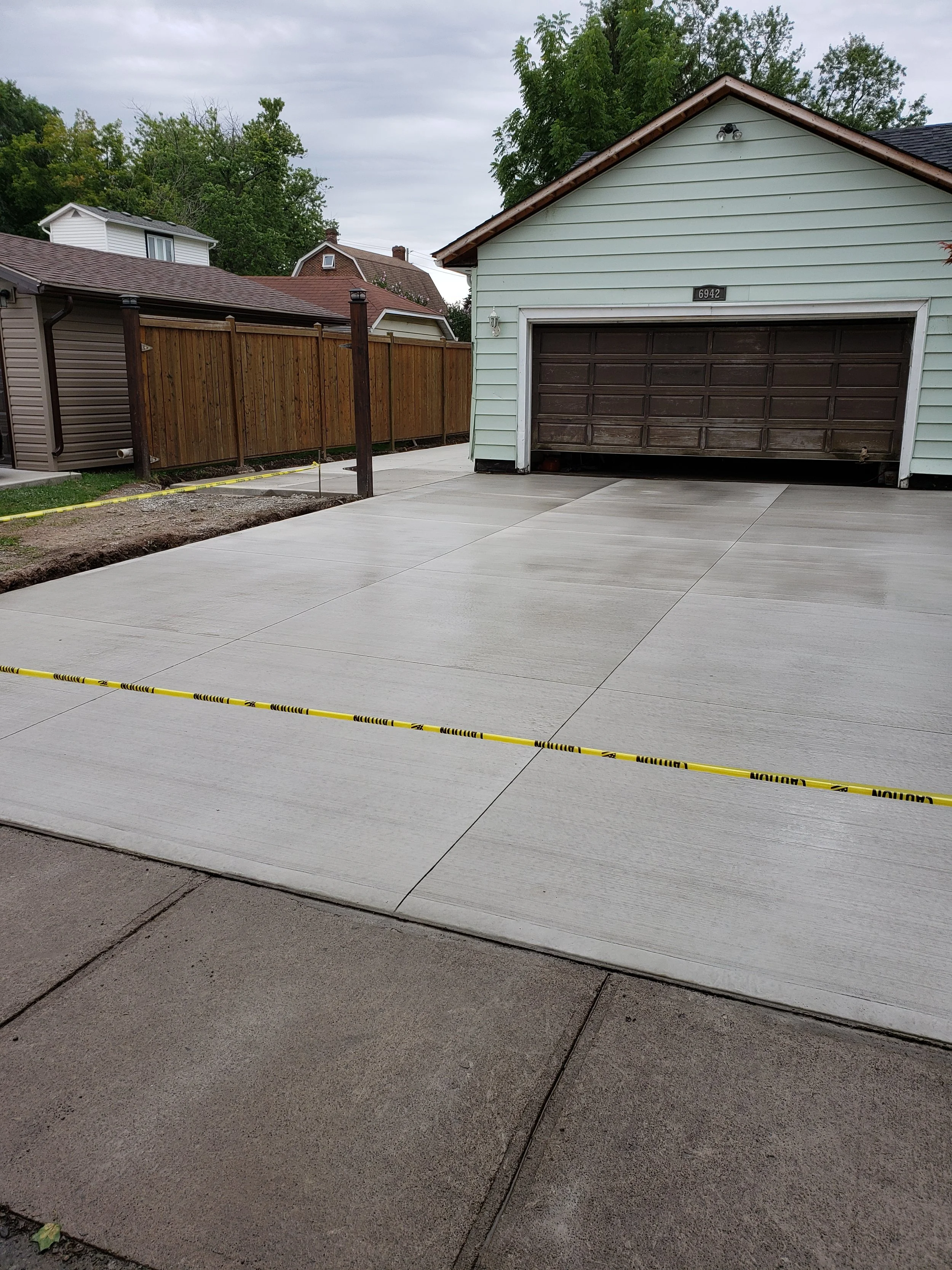Newly poured concrete driveway with yellow caution tape blocked off in front of a garage and a wooden fence, with neighboring houses and green trees in the background.
