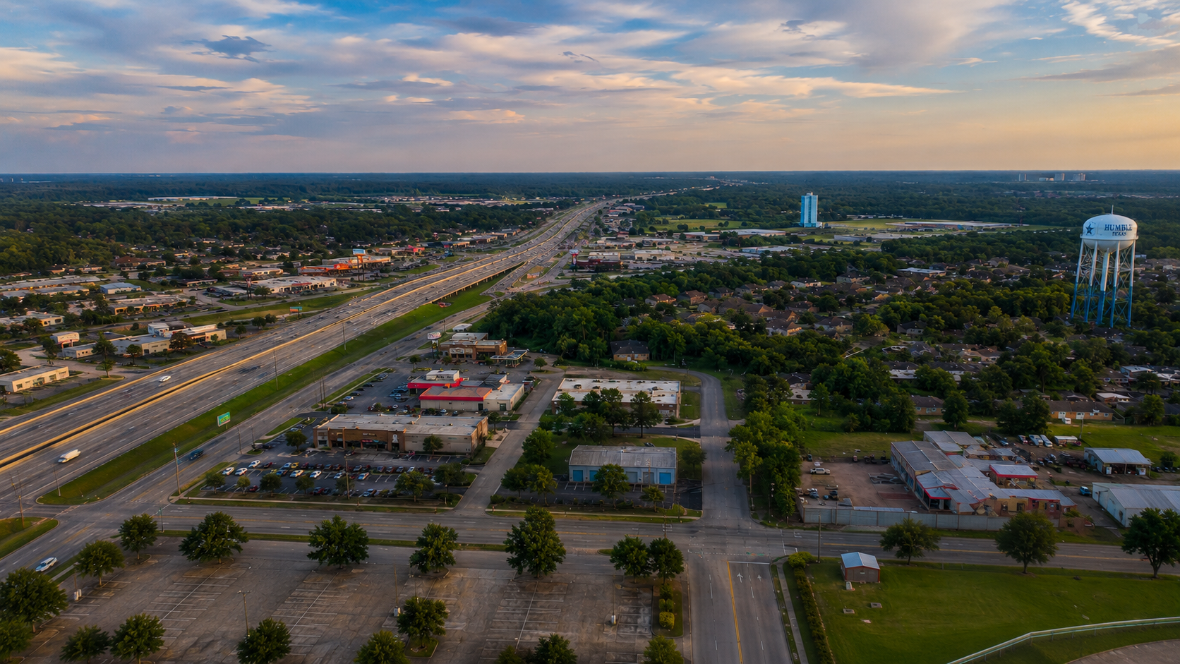 Aerial view of a suburban area with a highway running through it, a McDonald's restaurant, a water tower labeled "Humble Texas," and a mixture of commercial and residential buildings, with trees and green spaces, under a partly cloudy sky.