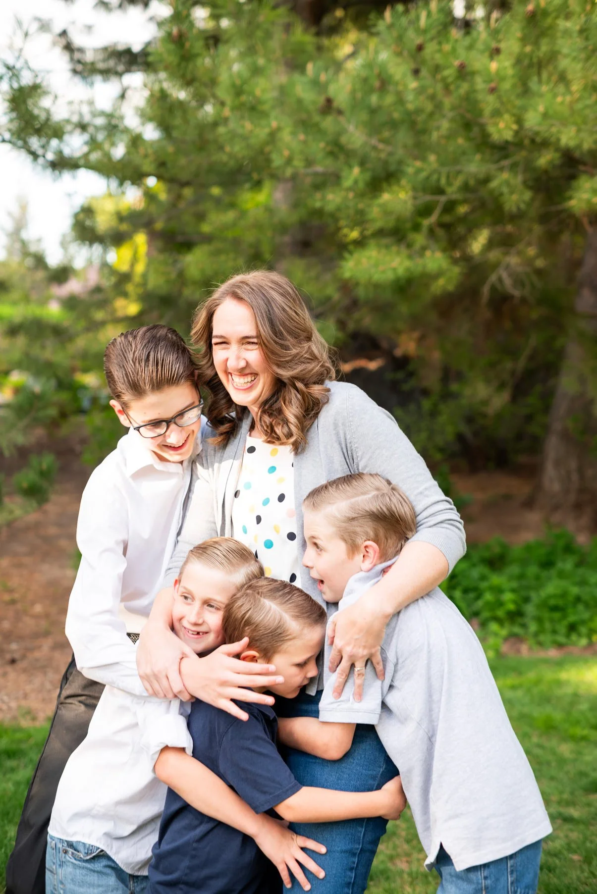 A woman and five children sharing a group hug outdoors in a park or garden with green trees in the background, all smiling and appearing happy.